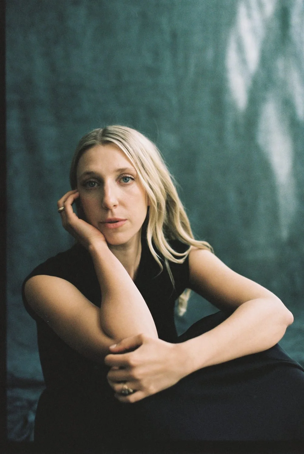 A woman with blonde hair and blue eyes wearing a black sleeveless top, sitting with her elbow resting on her knee, and her hand touching her face, against a dark green background.
