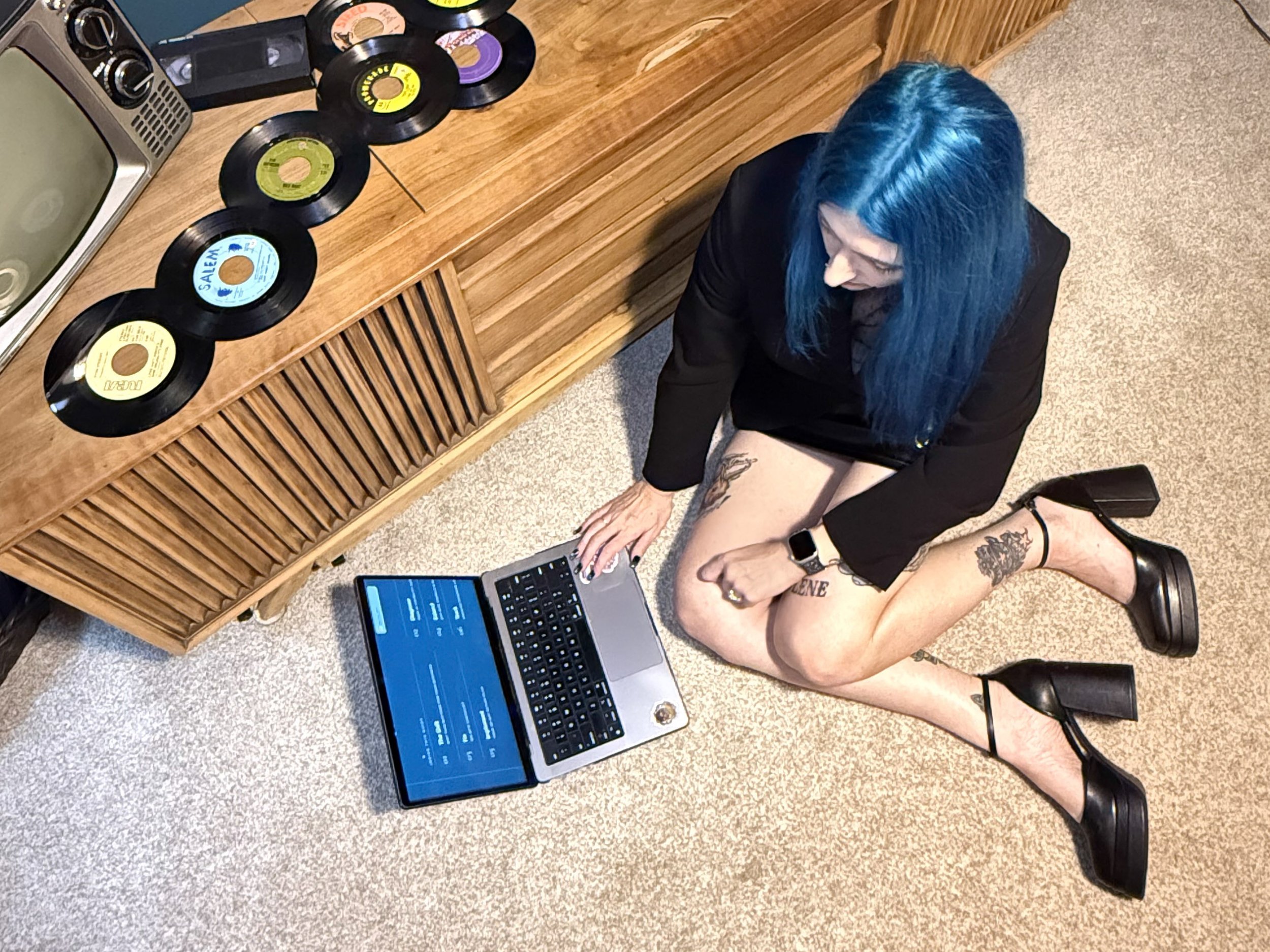 Woman with blue hair sitting on the floor using a laptop, with a keyboard and smartphone nearby, adjacent to a wooden cabinet with vinyl records, a small vintage TV, and an old radio on top.