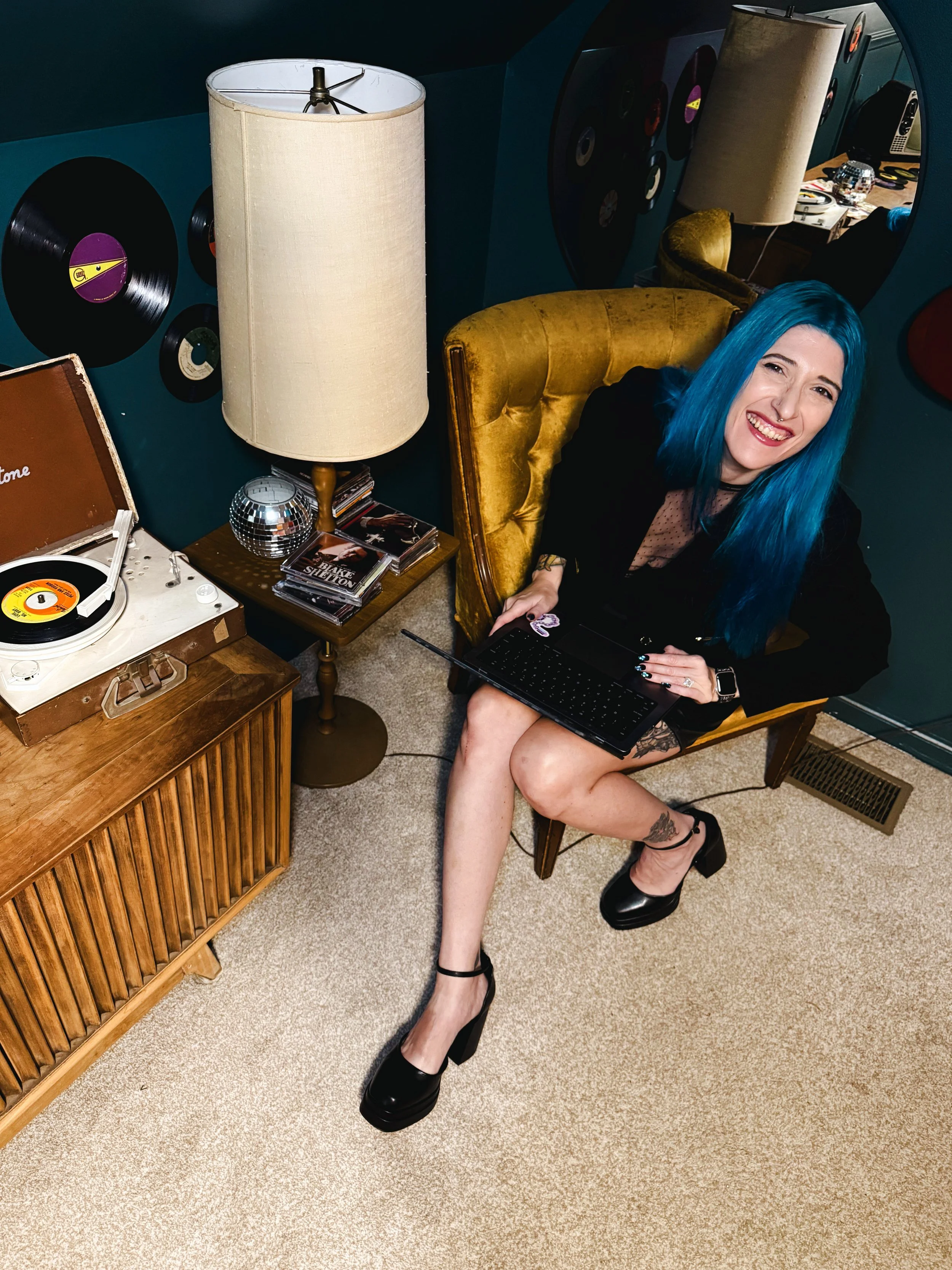 woman with bright blue hair seated on a yellow velvet armchair, smiling, with black high heels, patterned tattoo on her right leg, and holding a laptop, in a room with vintage decor and vinyl records on the wall.
