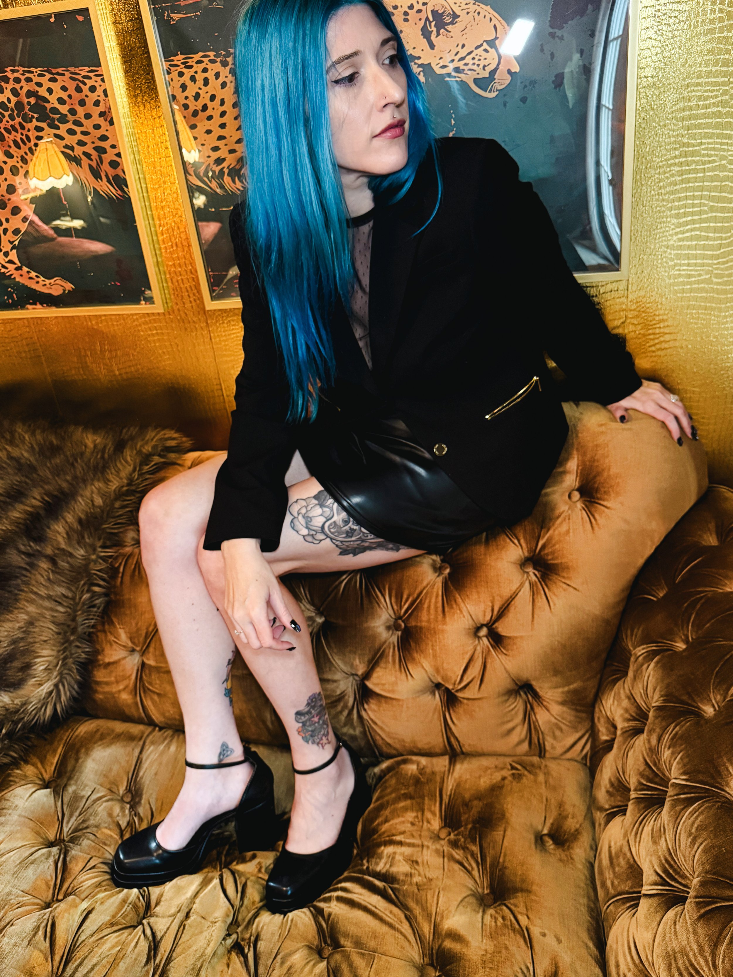 A woman with bright blue hair, wearing a black outfit, sitting on a vintage tufted leather couch with a leopard print. She has tattoos on her legs and is wearing black high heels. The background features artwork of a leopard and an orange and black color scheme.