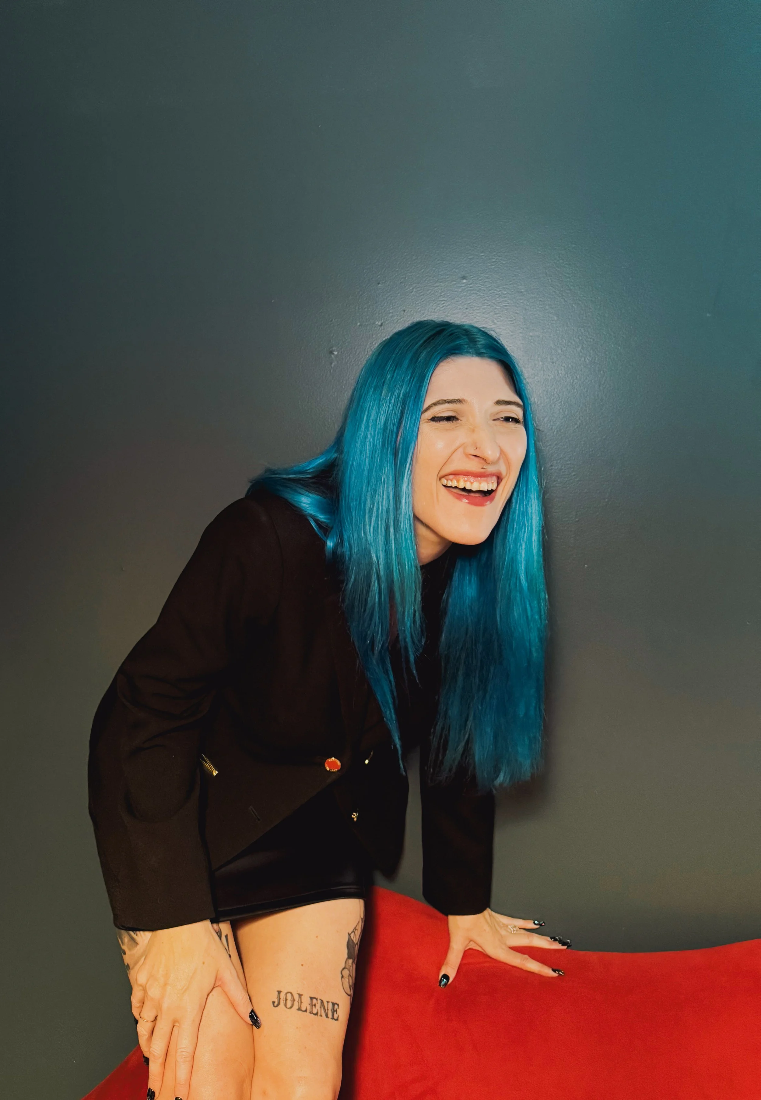 A woman with bright blue hair laughing, wearing a black blazer with gold buttons, and black nail polish. She has tattoos on her thighs, including the word 'JOLENE.' She is touching a red surface with her right hand, and her head is tilted back with a joyful expression.