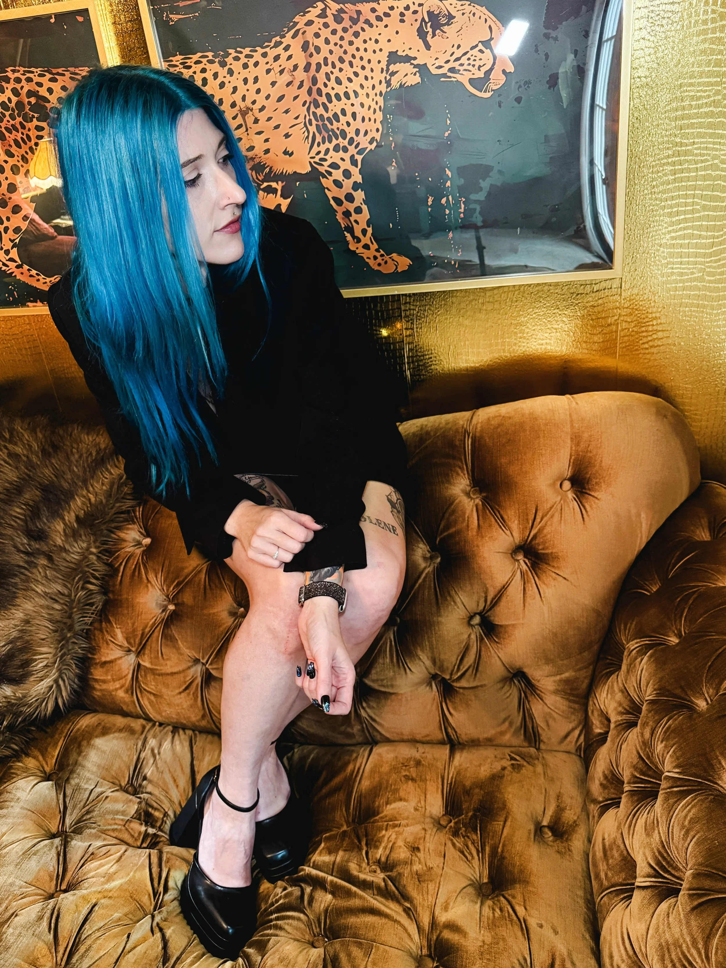 A woman with long blue hair sitting on a tufted leather sofa in a room with gold-colored walls and a large poster of a cheetah in the background.