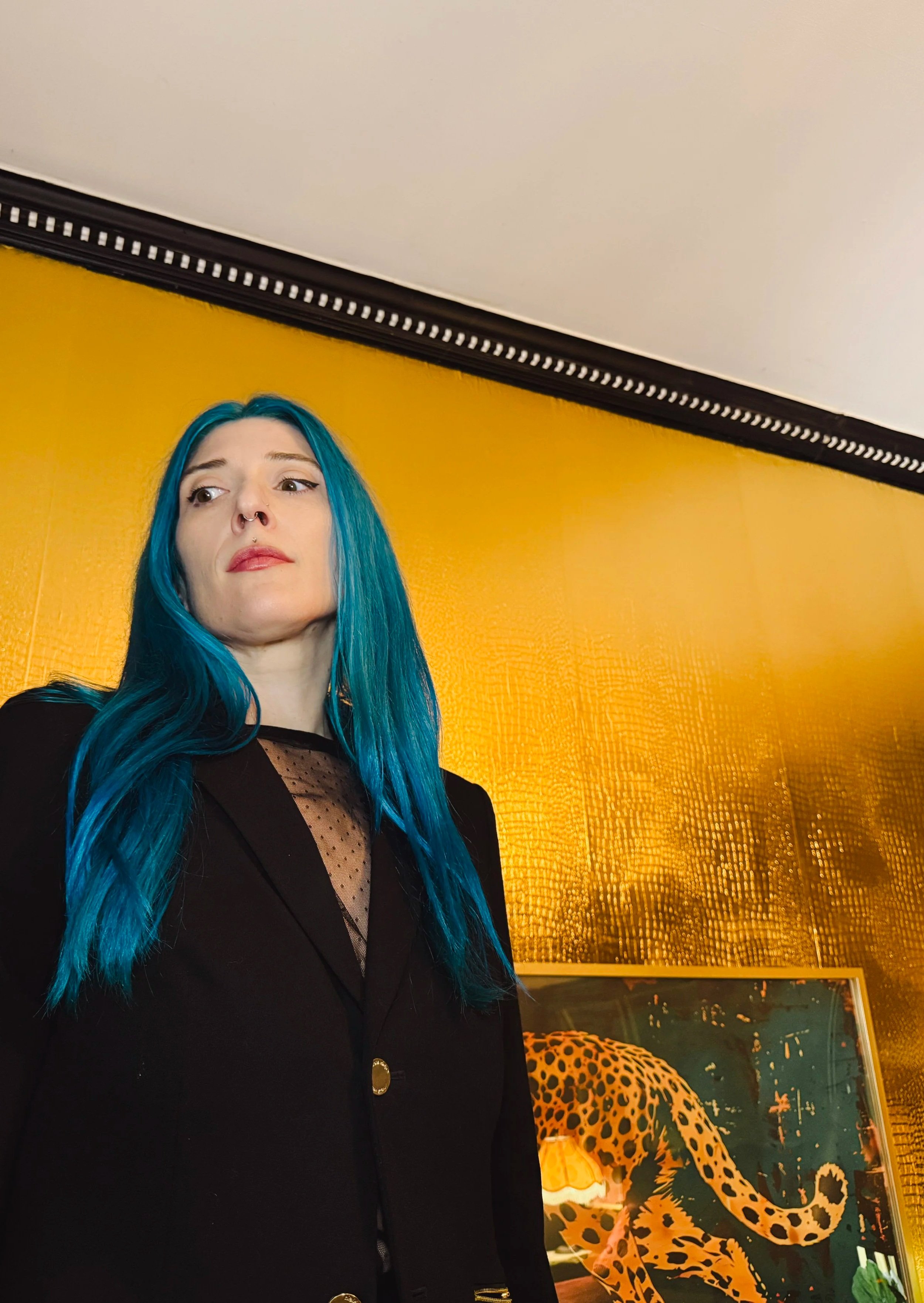 A woman with long blue hair and a septum piercing stands in front of a yellow textured wall with a leopard print sculpture and a painting of a leopard in the background.