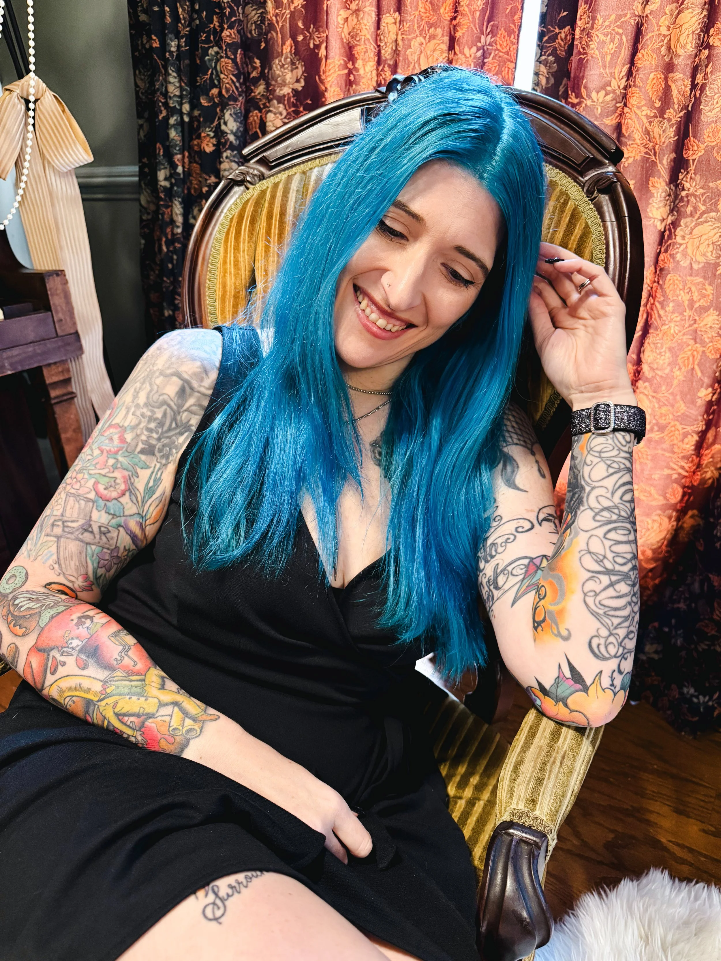 A woman with long bright blue hair smiling and sitting in a vintage armchair with a yellow and brown striped upholstery. She has tattoos on her arms and chest, is wearing a black sleeveless top, and has multiple rings and a black watch. Behind her are red floral curtains and a dark green wall. There's a beige pleated lampshade with a beaded string hanging beside her.