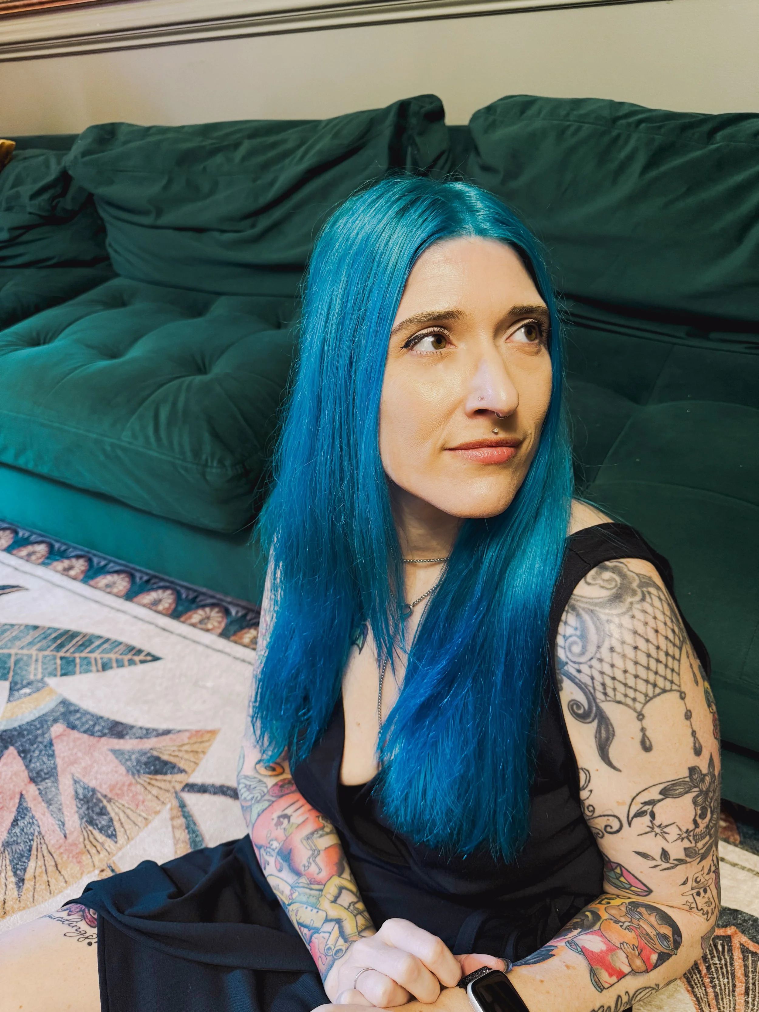 A woman with long bright blue hair sitting on the floor in front of a green couch, showcasing her tattoos on her arms and legs. She is wearing a black sleeveless top and has multiple tattoos visible on her arms and legs.