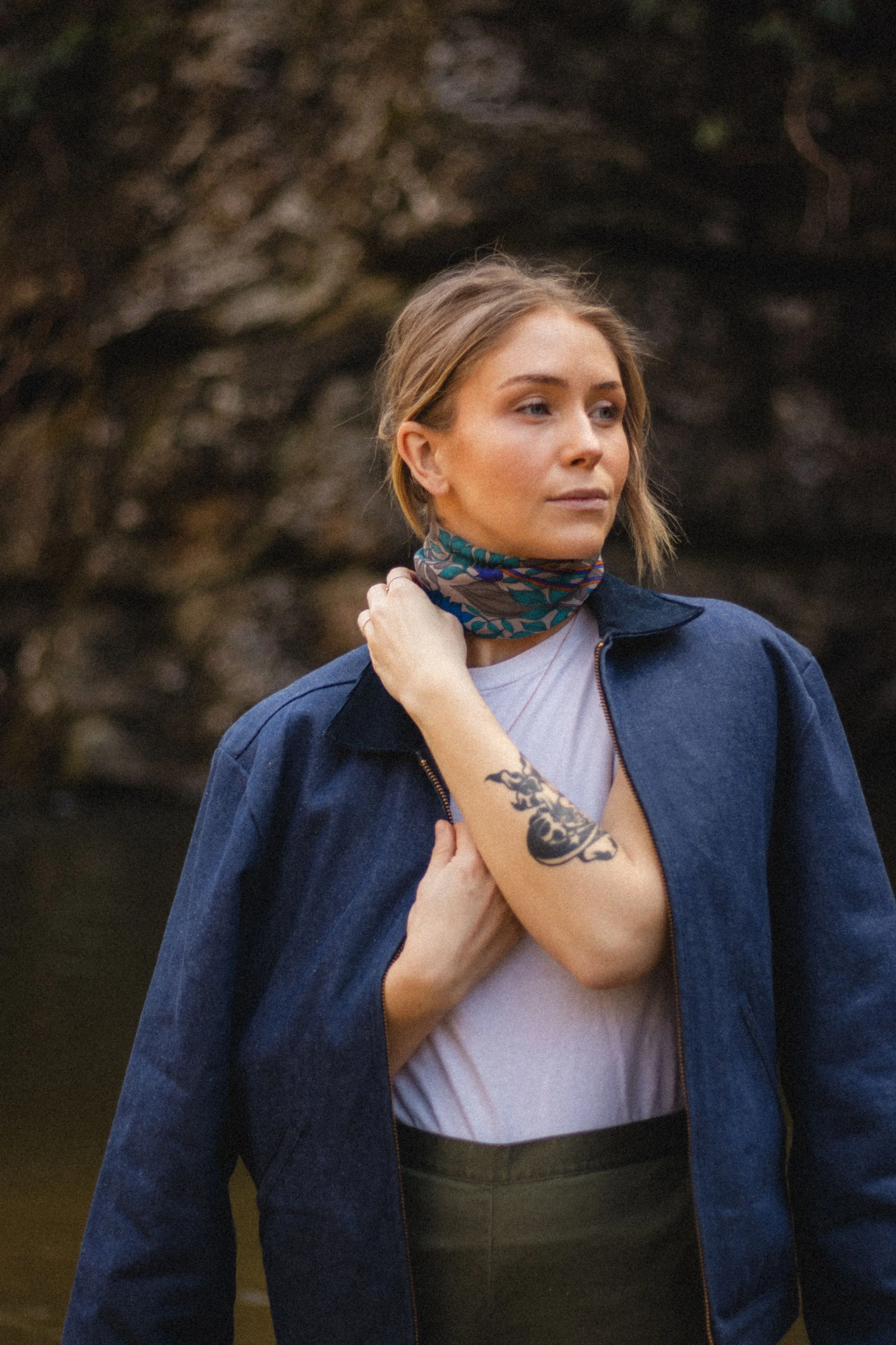 Portrait of a woman wearing a navy jacket and patterned neck scarf, standing outdoors against a dark rocky background with a tattoo visible on her forearm.