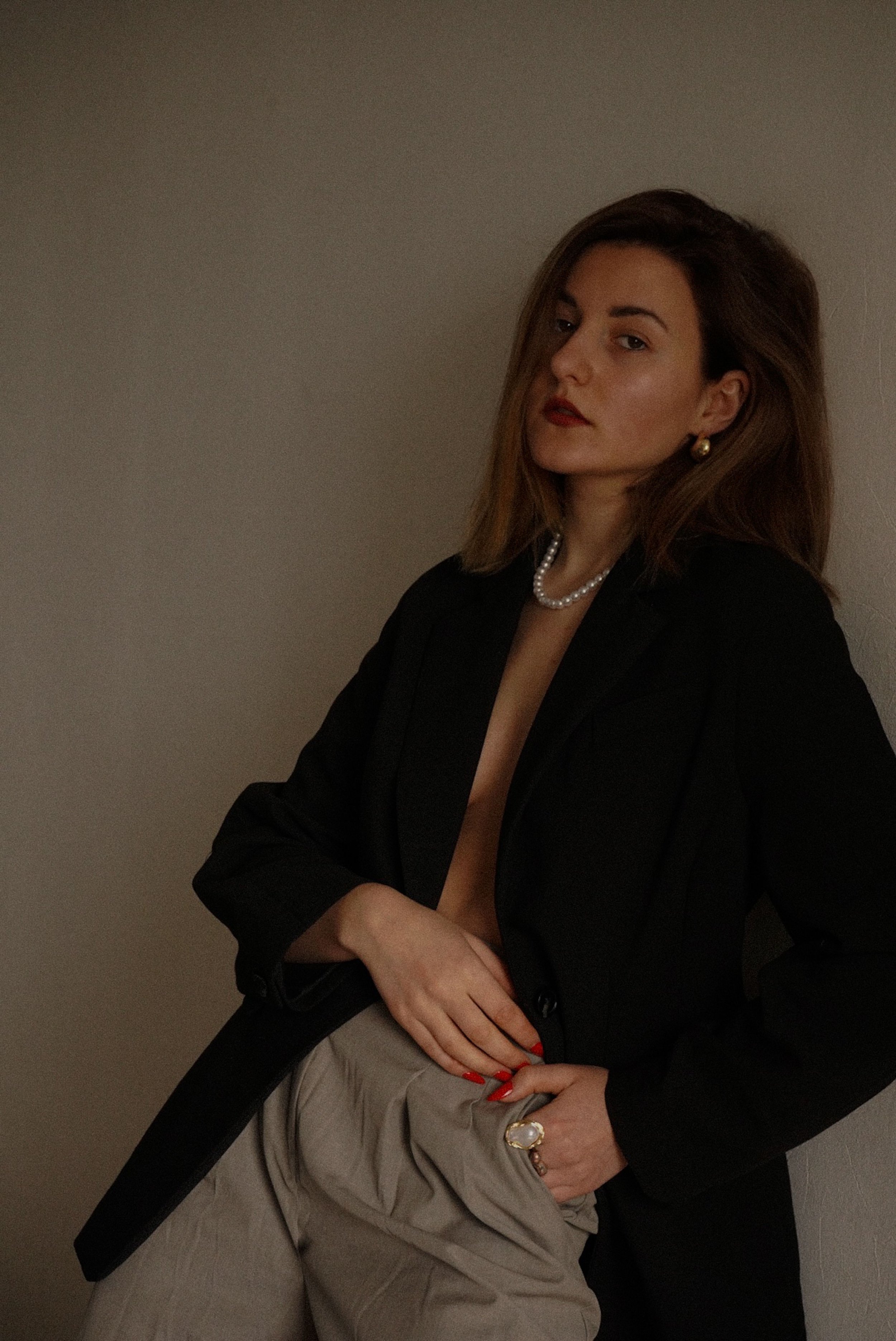 Editorial portrait of a woman in a black blazer, pearl necklace, gray trousers, and red nails leaning against a neutral wall and looking toward the camera.