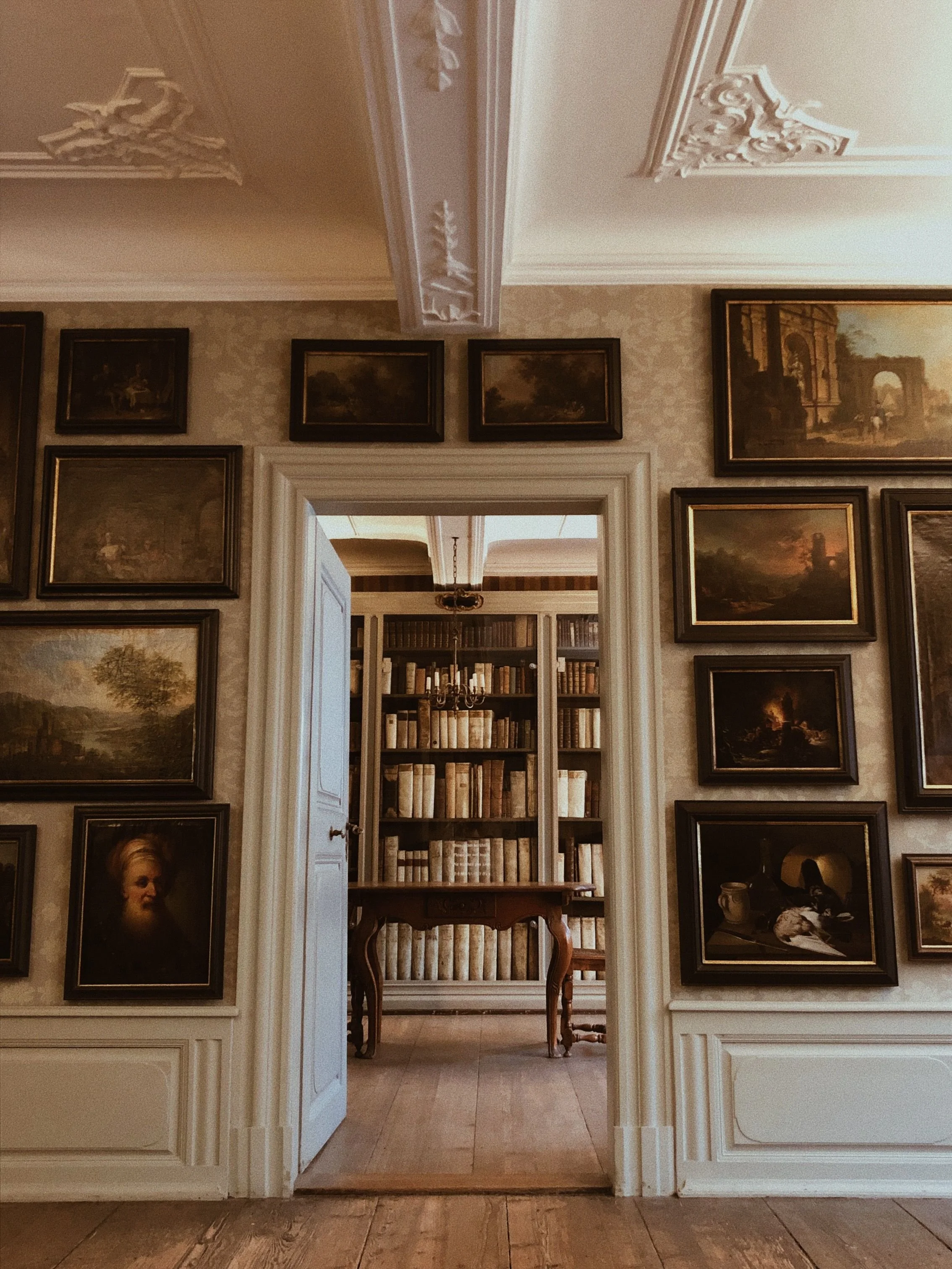 Open doorway leading into a library, surrounded by framed paintings in a warm, historic interior.