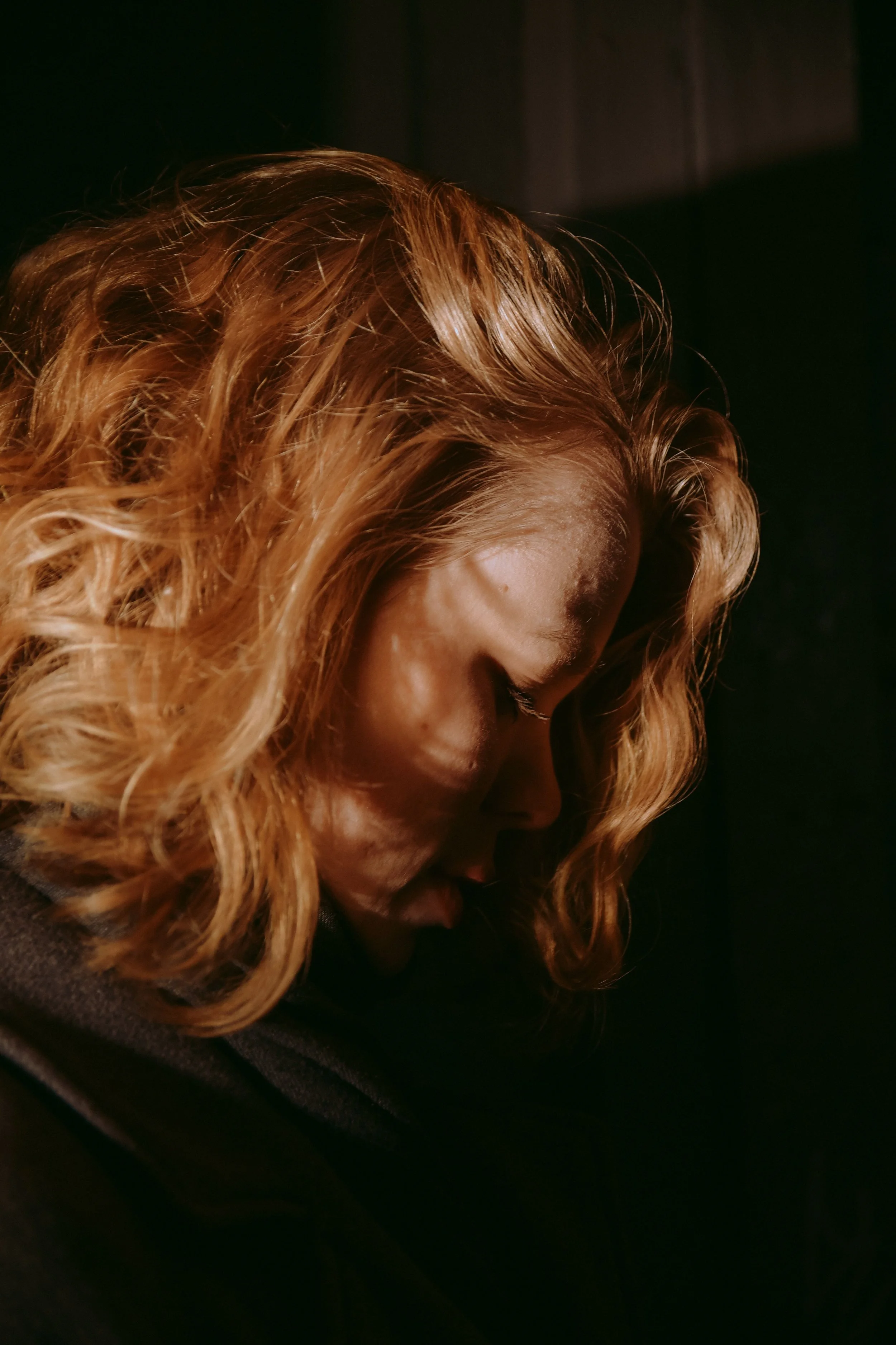 Woman with wavy blonde hair looking downward in warm amber light, with soft shadow patterns falling across her face against a dark background.