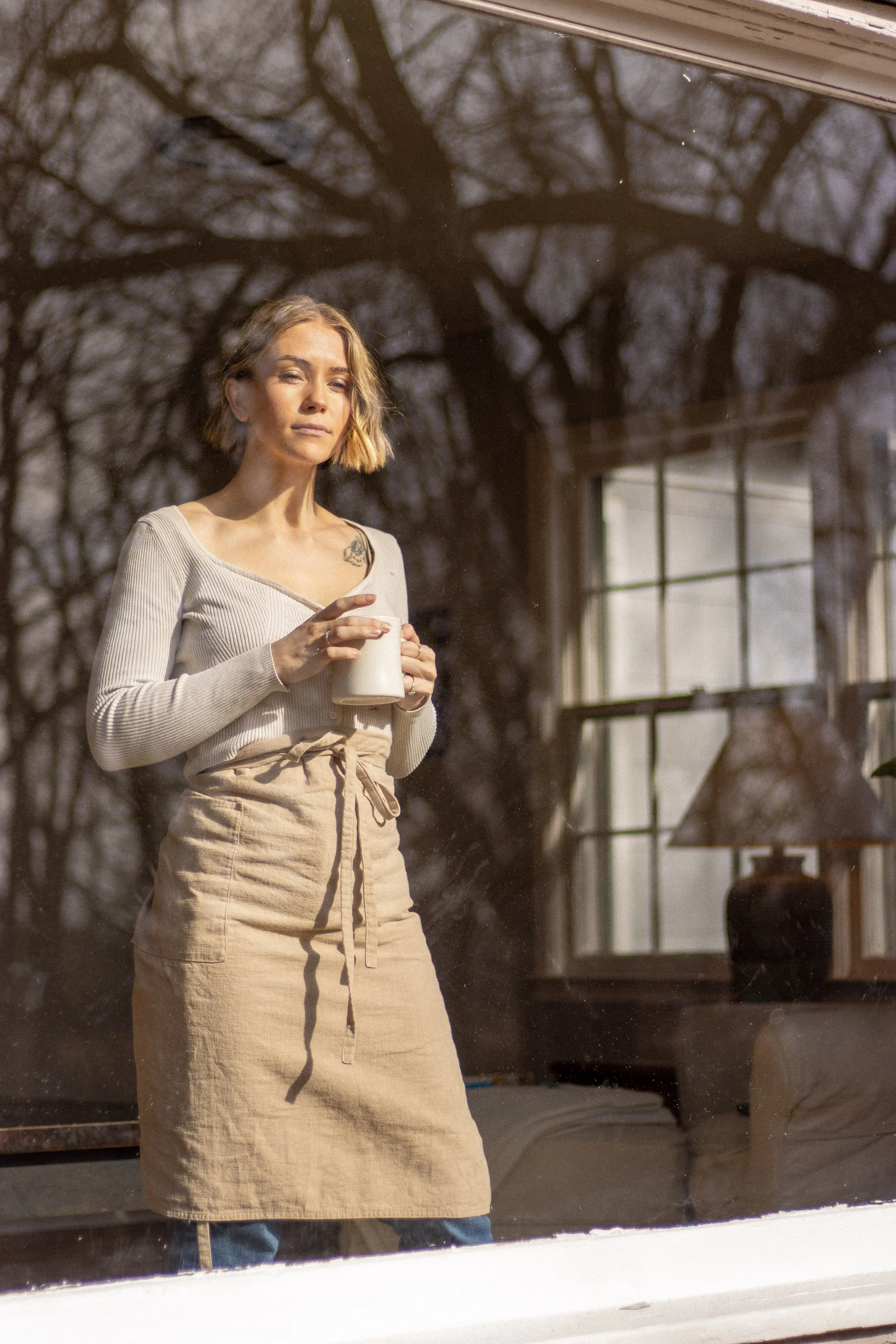 A woman standing indoors in front of a window, holding a mug, with a shadow of leafless trees visible through the window.