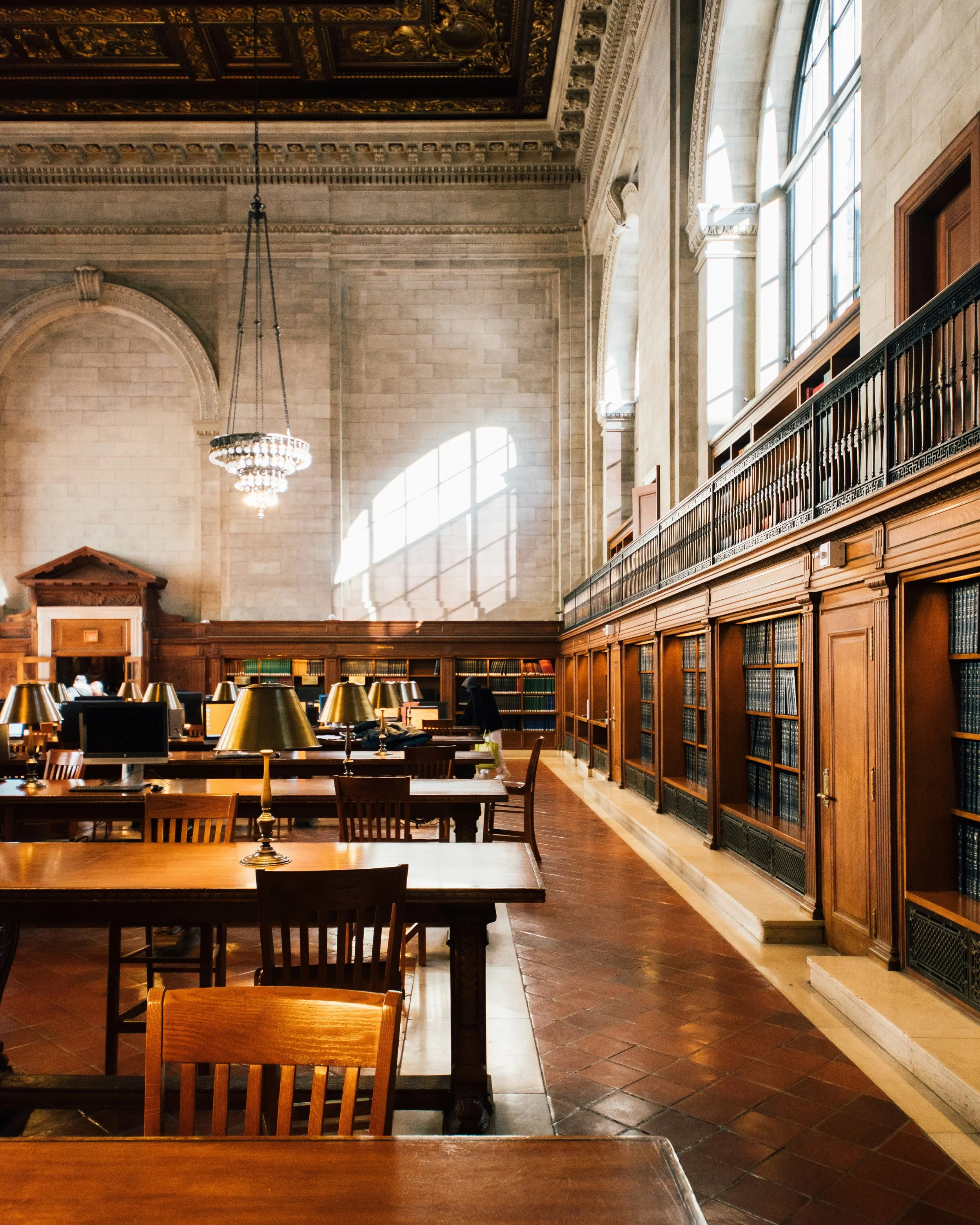 Grand library reading room with long wooden tables, bookshelves, and natural light.