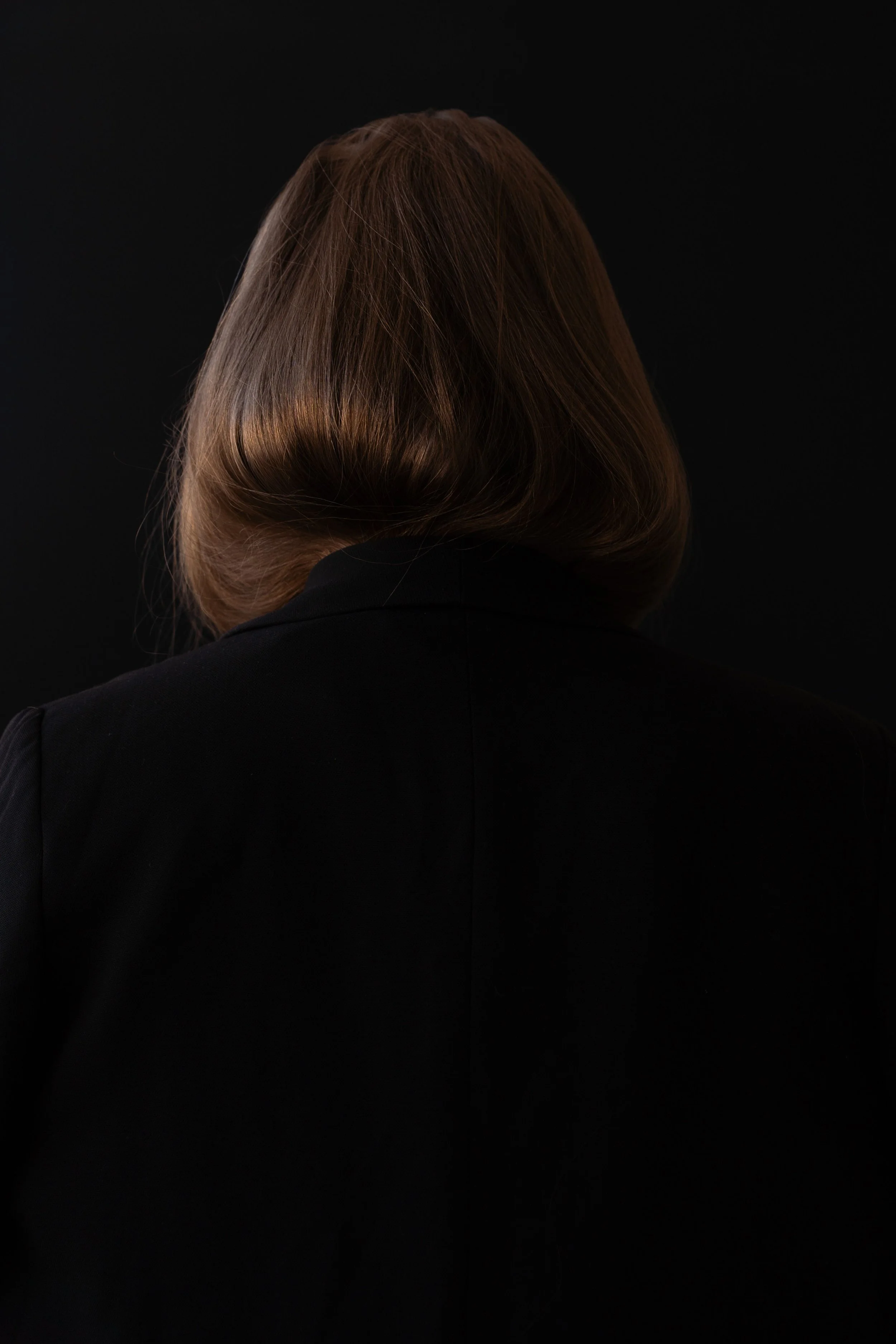 Back view of a woman in a black blazer with a smooth brown bob haircut, centered against a solid black background.