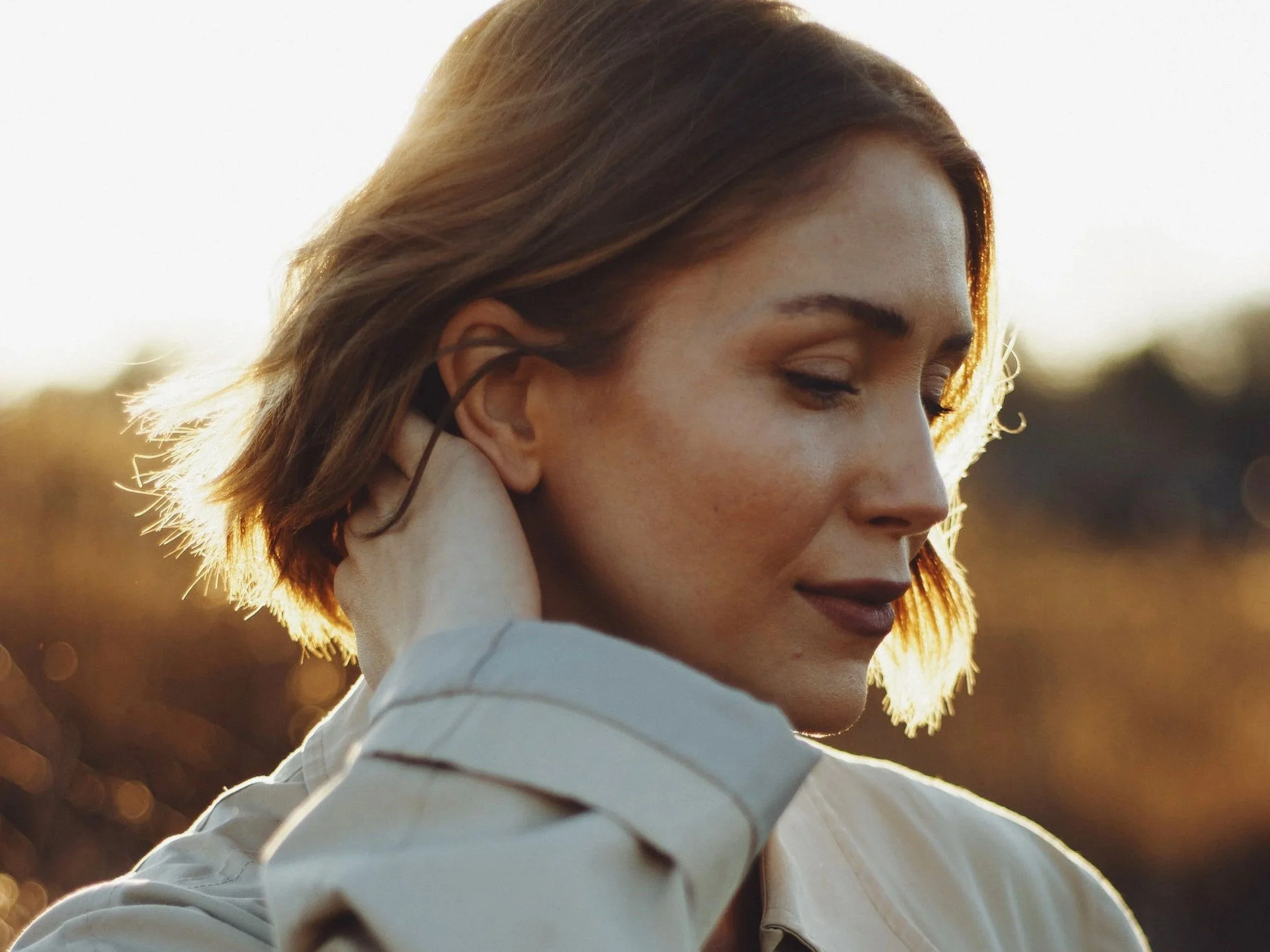 Portrait of a woman in a beige trench coat outdoors in warm golden-hour light, looking downward with one hand resting at the back of her neck.