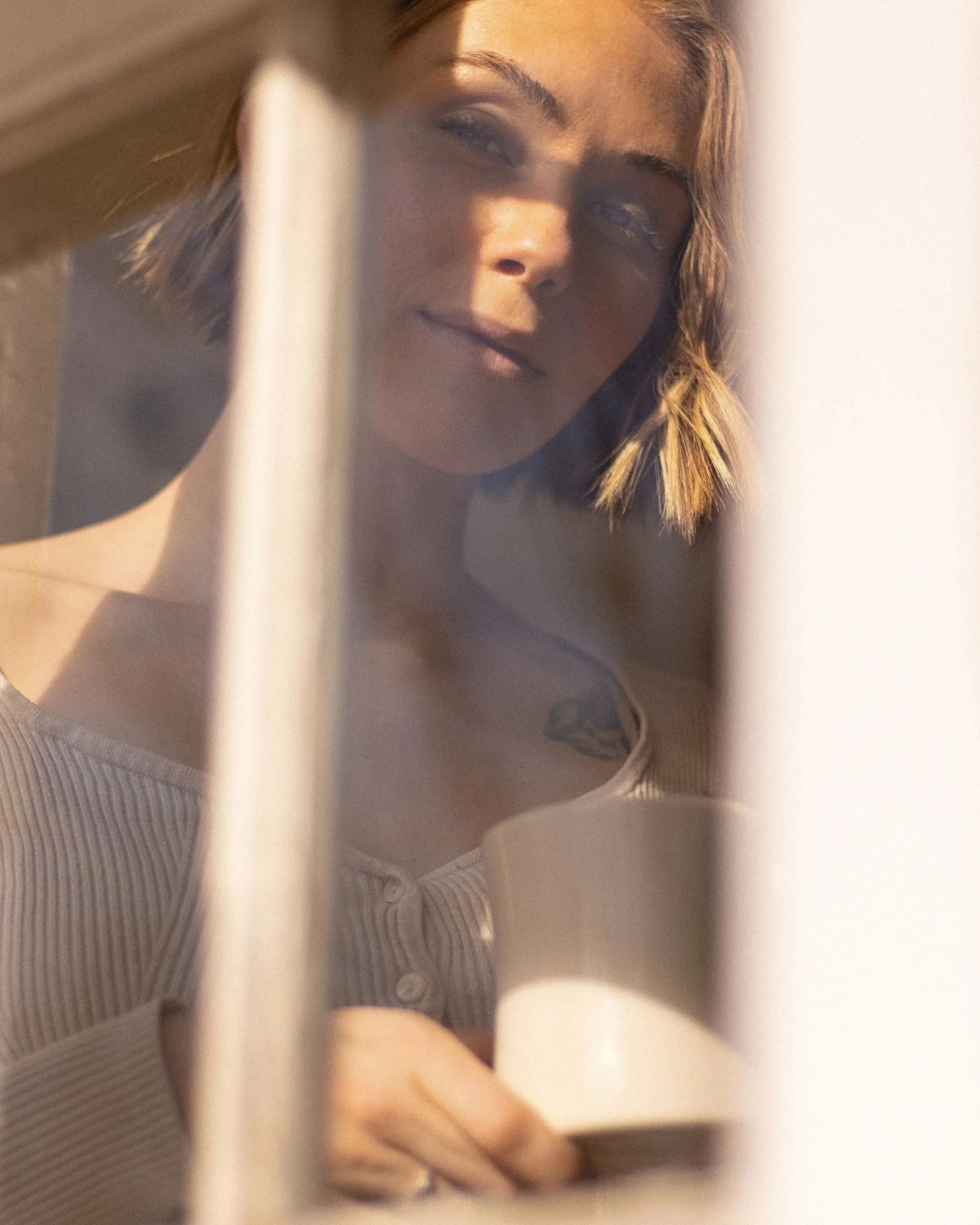 Woman framed by soft architectural lines, looking through a sunlit interior with warm shadows across her face.