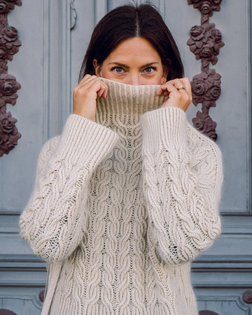 Woman with dark brown hair and blue eyes wearing a cream-colored knit sweater pulling it up to cover her lower face, standing in front of a decorated door with floral accents.