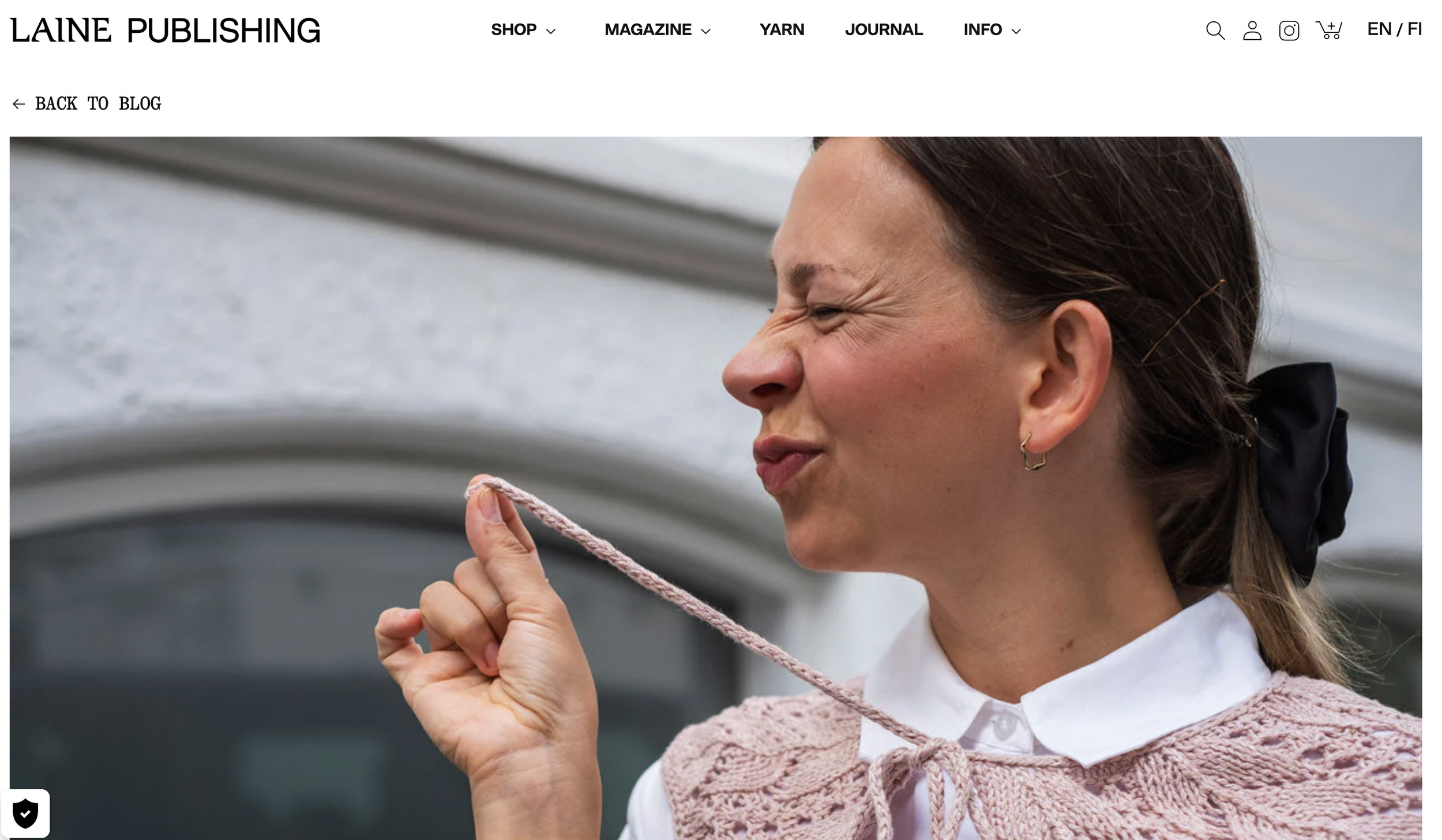 Woman making a face of disgust while holding and smelling a piece of pink yarn, standing outdoors in front of a building.