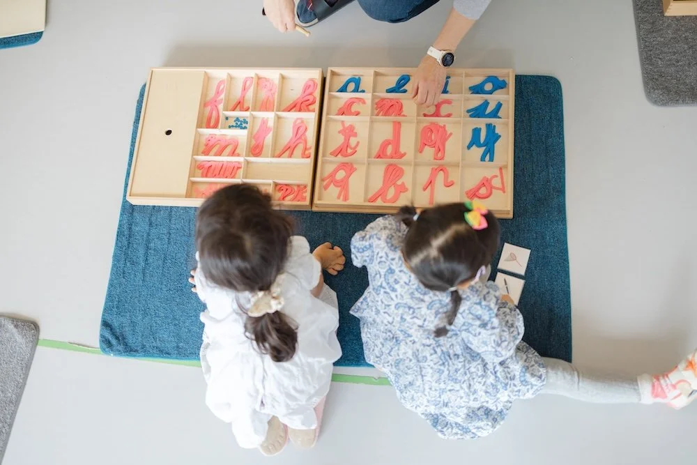 Deux enfants jouent avec des lettres cursives en plastique rose et bleu sur une table en bois. Un adulte supervise la activité.