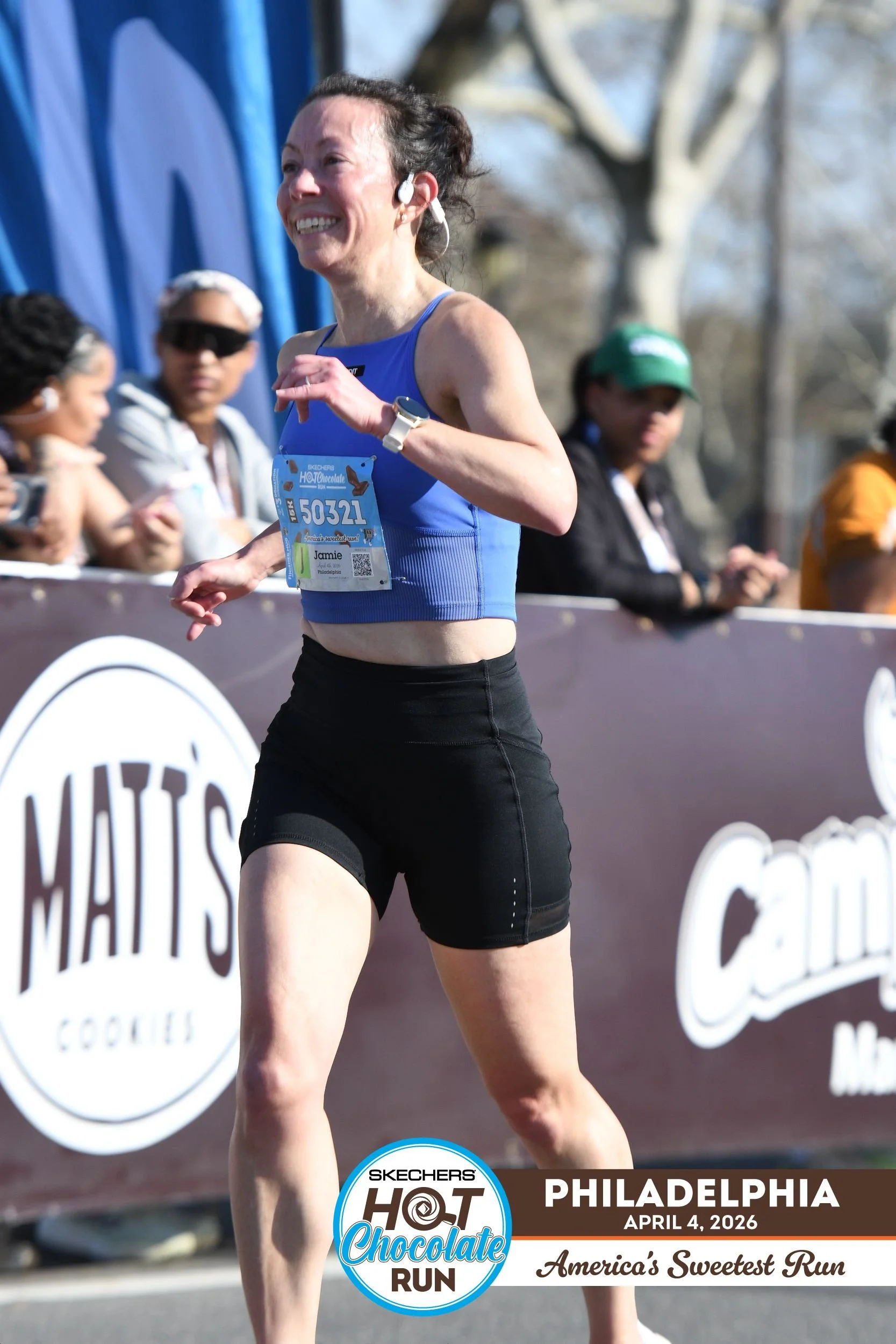 A woman running in a marathon, wearing a blue sports top and black shorts, with a race bib numbered 50321. She is smiling and appears to be enjoying the race on a sunny day. There are spectators and other runners along the course, and banners with logos, including "Matis Cookies" and "Calloway" in the background. The event is the Skechers Hot Chocolate Run in Philadelphia, held on April 4, 2026, and is described as "America's Sweetest Run."