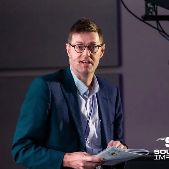 A man with glasses and short dark hair, wearing a dark blue blazer and light blue shirt, holding papers in his hand, standing in front of a dark background.