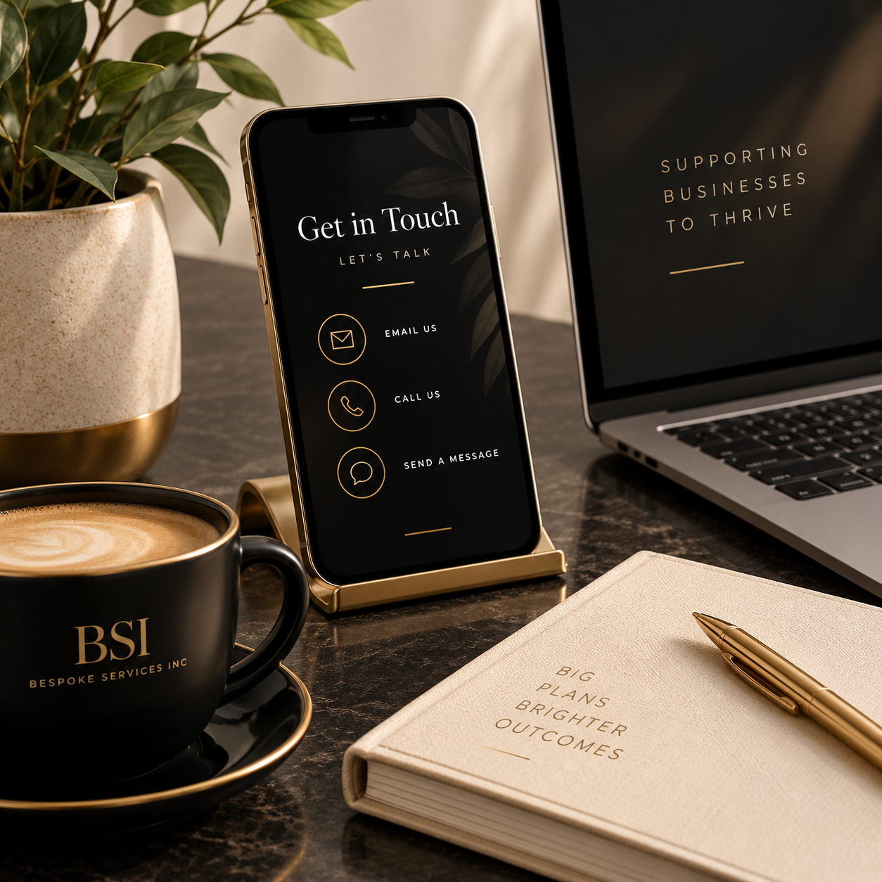 A desk setup with a potted plant, a cup of coffee labeled 'BSI BESPOKE SERVICES INC', a smartphone on a stand displaying contact options, a closed notebook titled 'BIG PLANS BRIGHTER OUTCOMES' with a gold pen, an open laptop, and part of a dark surface.
