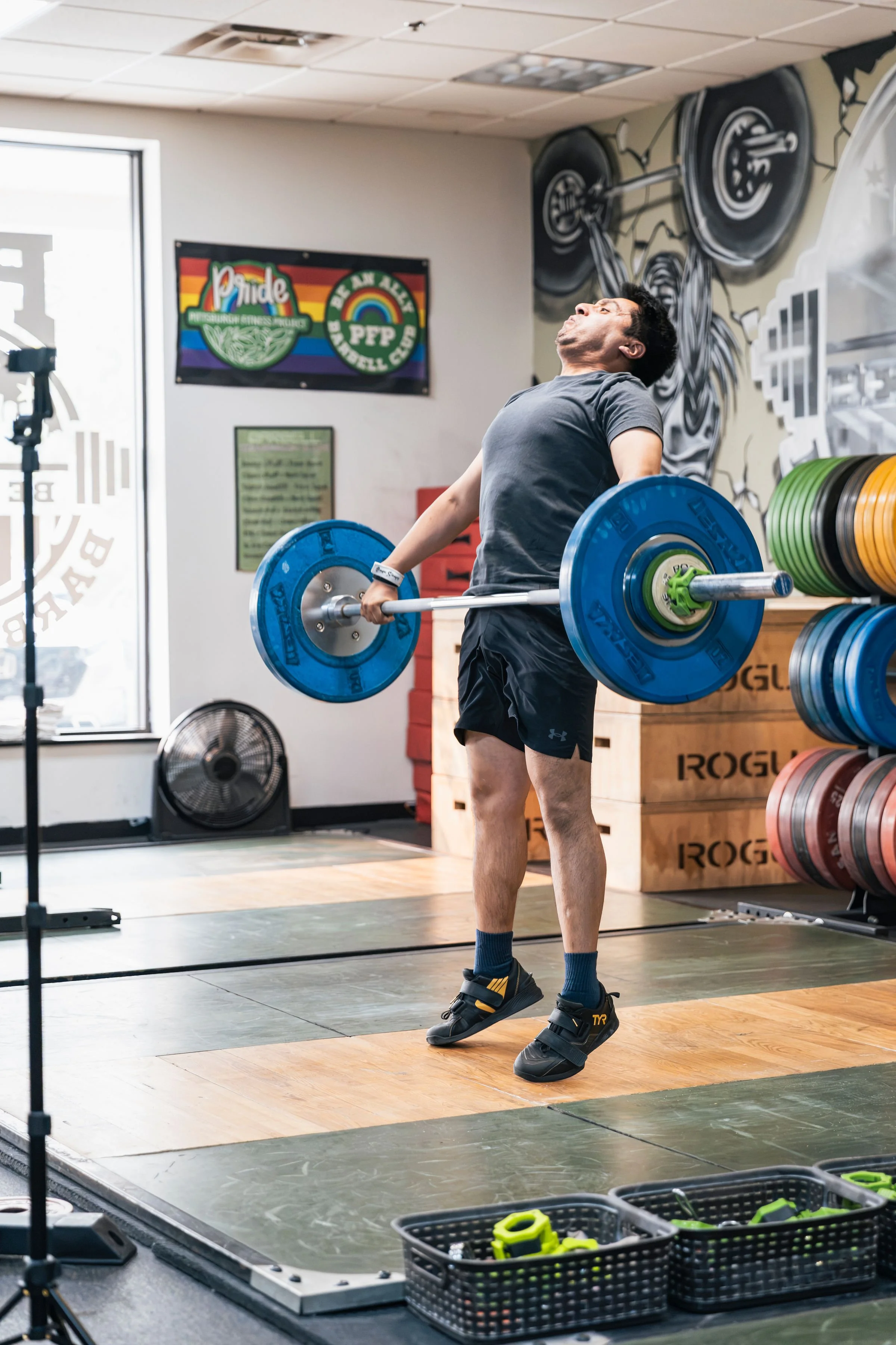 A man lifting a barbell with blue and green weight plates in a gym. The gym has a mural of weights on the wall and pride flags. There are storage shelves with additional weights and gym equipment, and a fan on the floor.