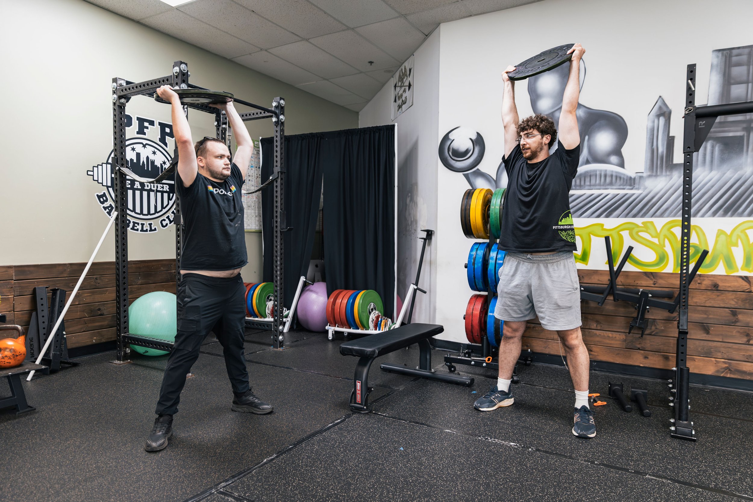 Coach Issaac working with a client lifting weight plates overhead in a gym, one on each side of a power rack, with various exercise equipment around them.