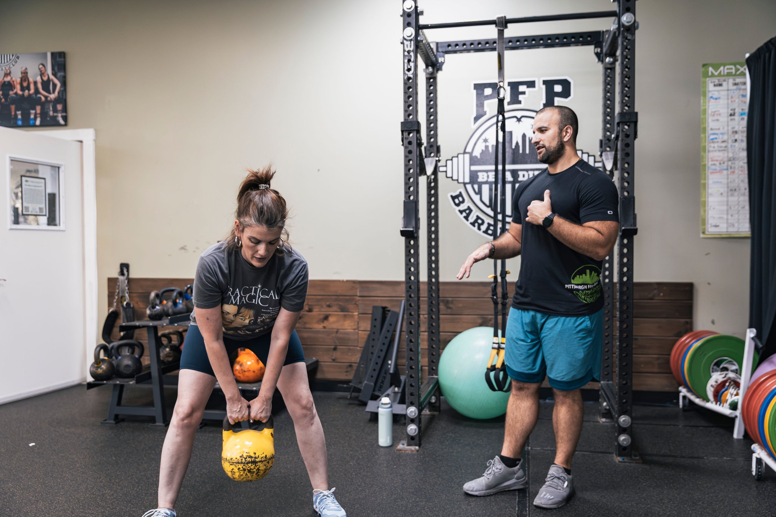 A woman is performing a kettlebell swing exercise in a gym, while Coach Frank watches her and gives instructions. The gym has various weightlifting equipment, including kettlebells, weight plates, and a power rack with a logo that reads 'PFP'.