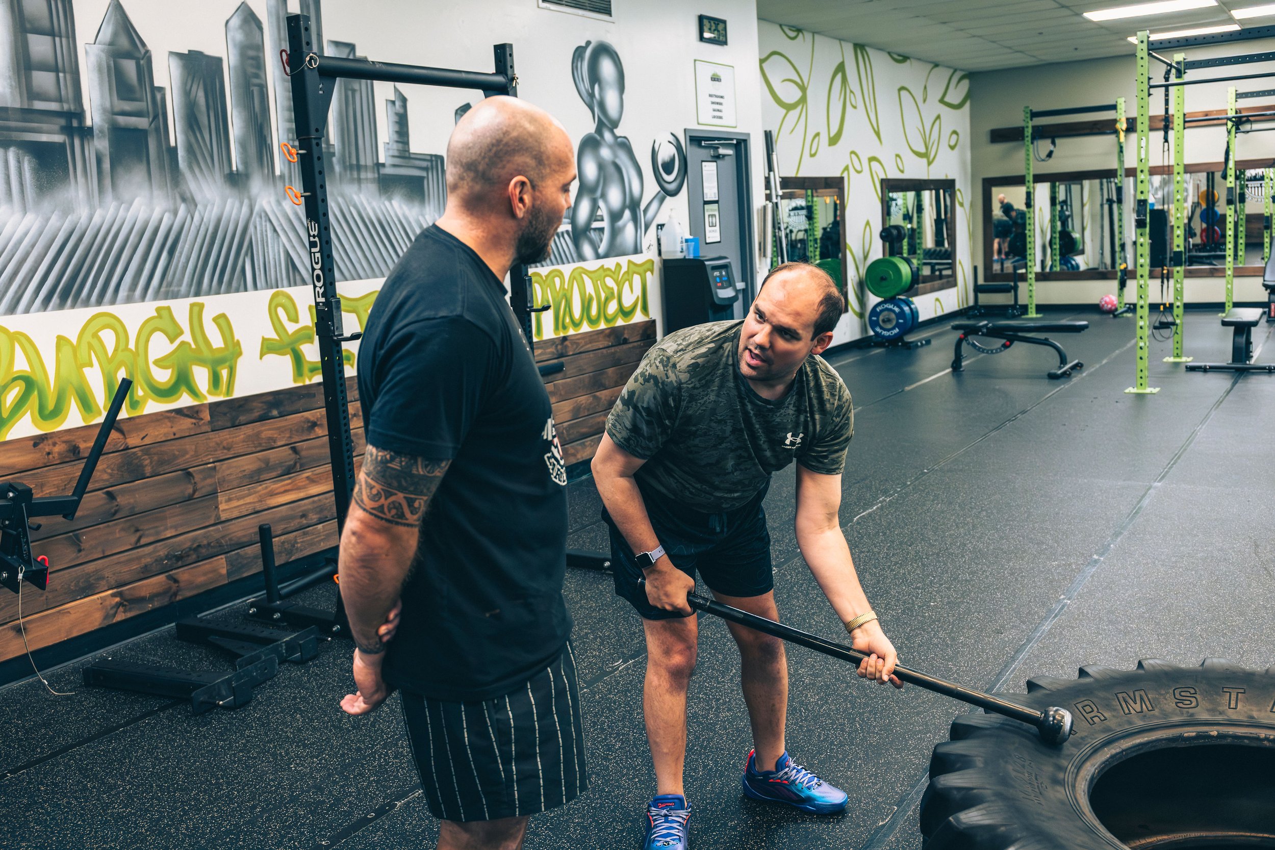Coach Wes assisting his client with a tire flip exercise, in front of gym equipment and wall art.