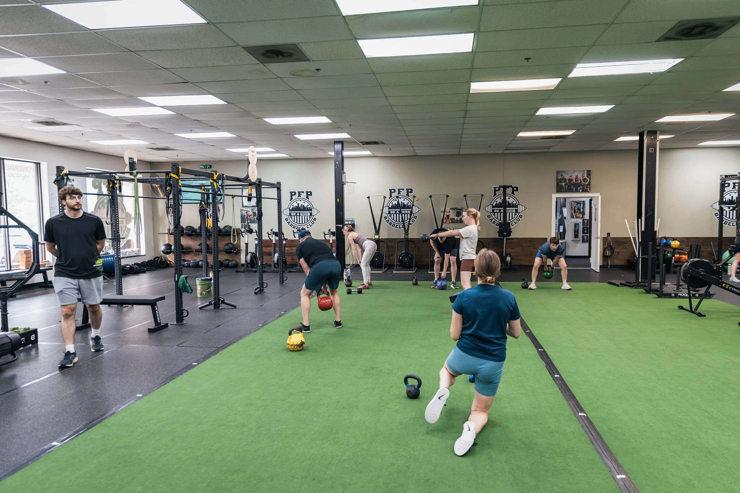 People exercising in a gym with various fitness equipment, including kettlebells, barbells, and pull-up bars, with a trainer and a group of participants.