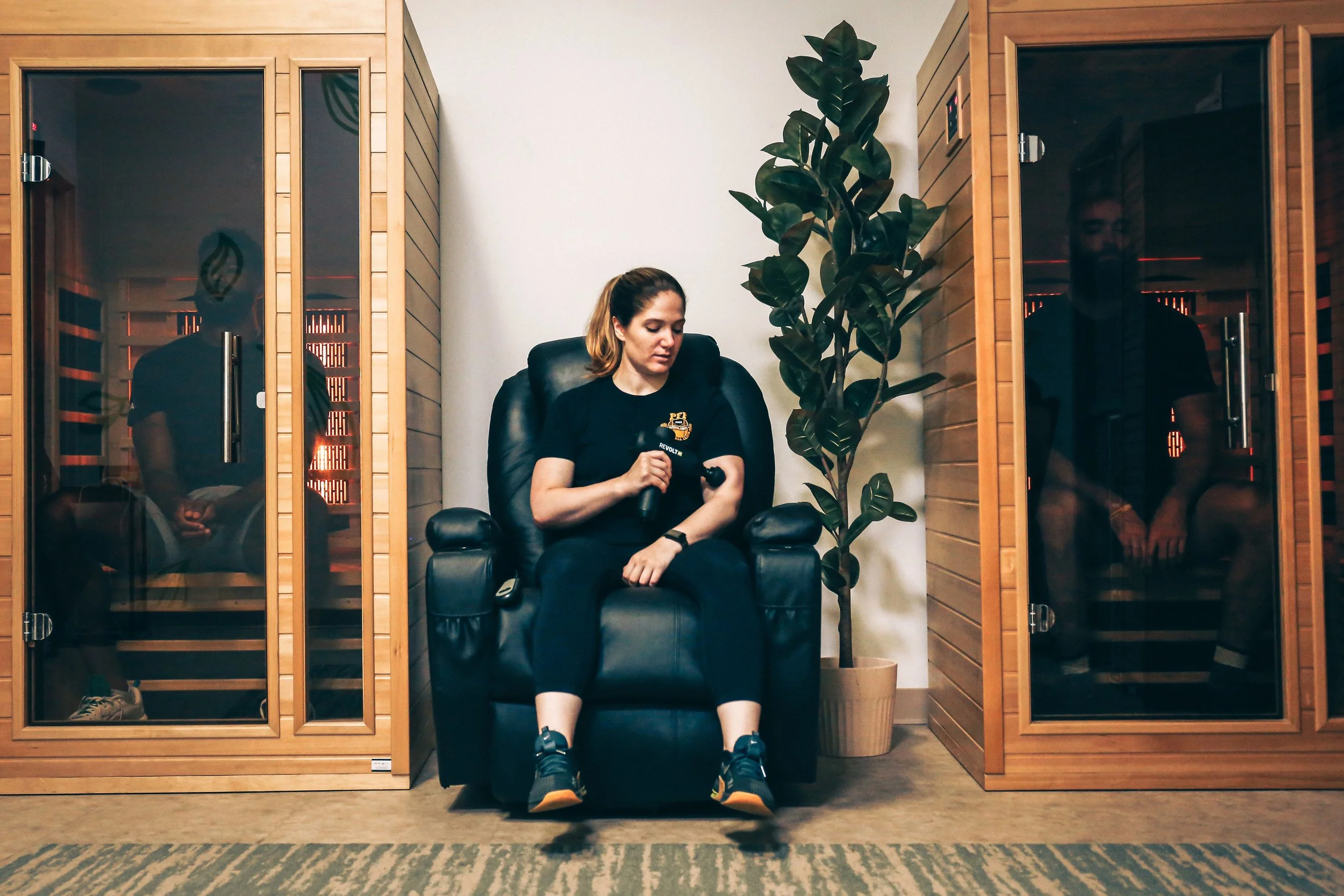 Coach Gina, sitting in a black leather chair inside a spa room. Behind her are two of PFP's wooden sauna cabins with tinted glass doors. (Other PFP trainers are barely visable through the tinted glass).