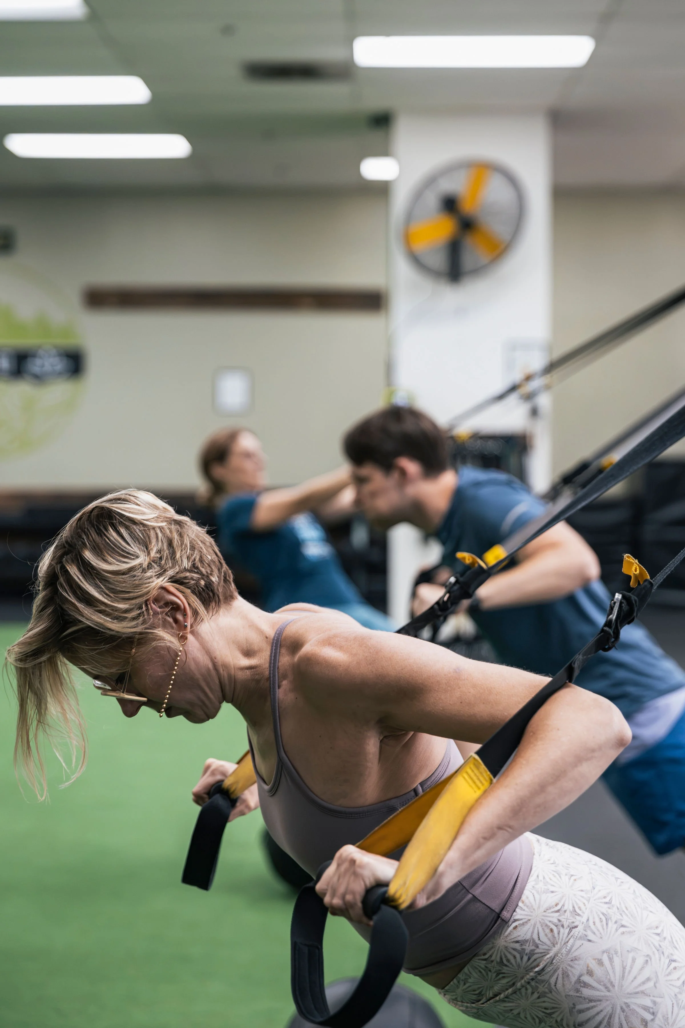 People doing suspension training exercises at a gym, with one woman in the foreground and others in the background, a clock on the wall and gym equipment visible.