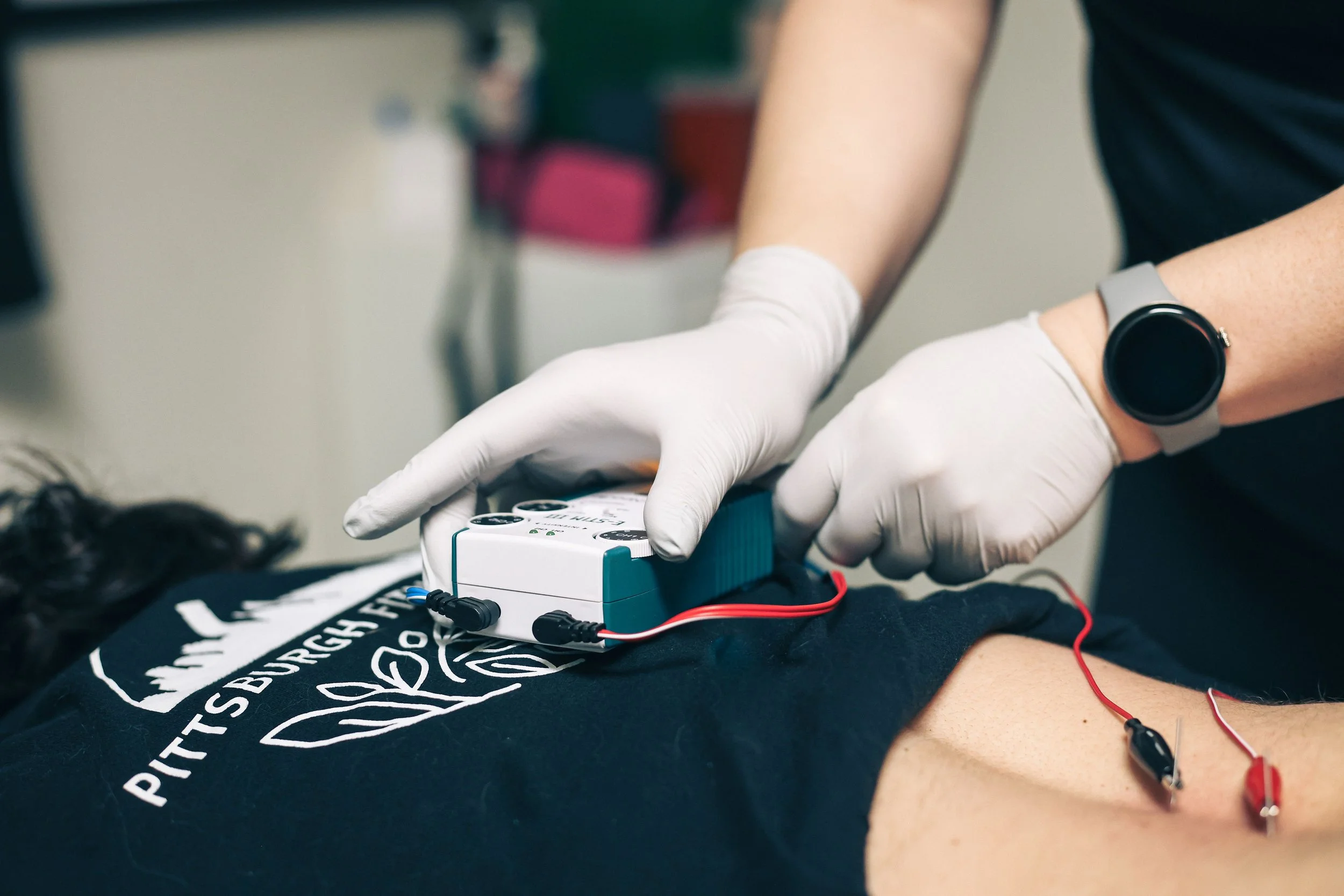 Person wearing a smartwatch and white gloves inserting an electronic device with wires into a person’s arm during a medical procedure.