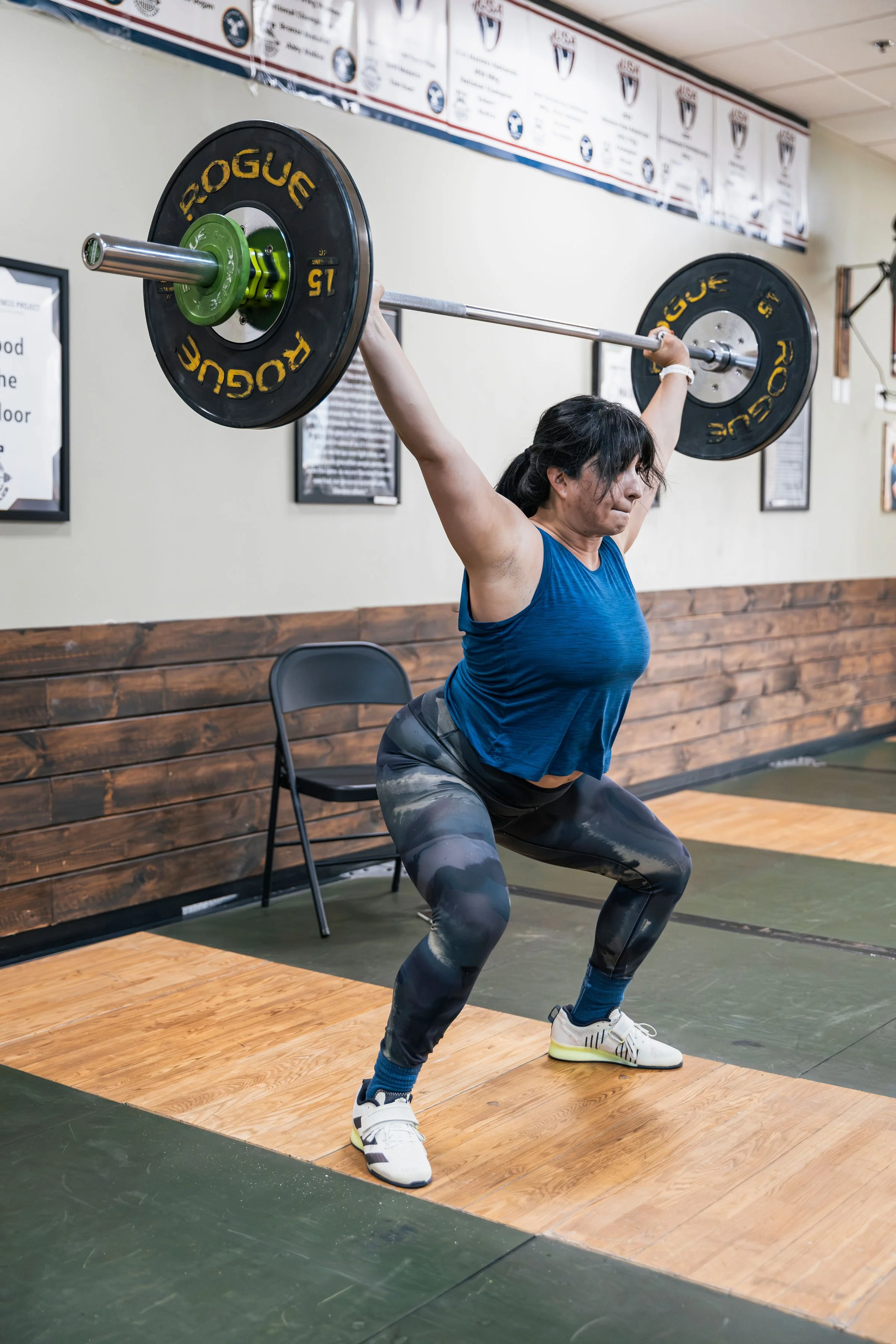 Woman lifting a barbell in a gym, wearing a blue tank top, camouflage leggings, and white sneakers, with a wooden floor and a wall with banners in the background.