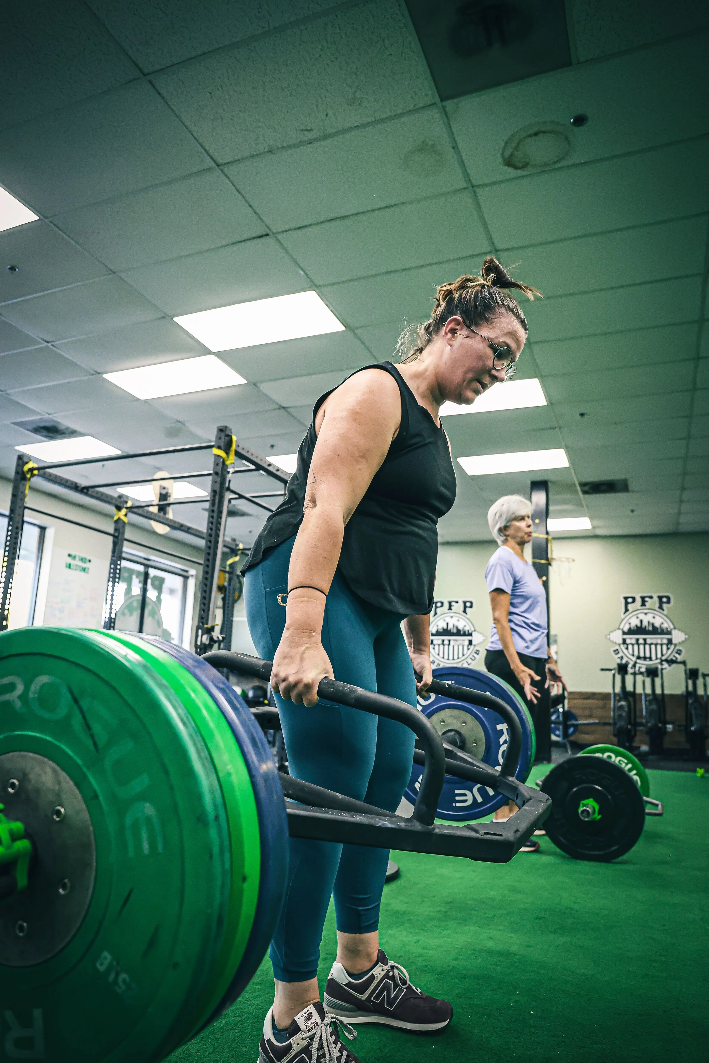A woman with glasses and a bun hairstyle, dressed in a black sleeveless top and blue leggings, is lifting a barbell with green weights in a gym. There is another woman in the background, wearing a purple shirt and white hair, also exercising. The gym has green flooring, black and yellow equipment, and a logo with the letters 'PFP' on the wall.