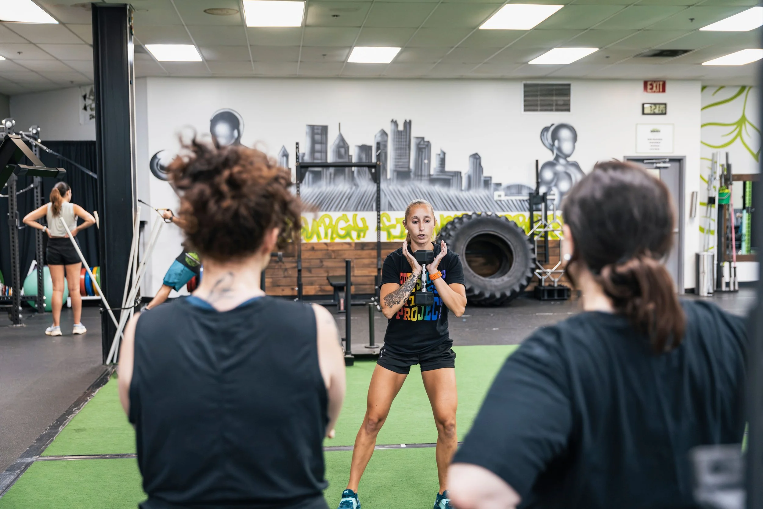 A fitness instructor leading a workout class in a gym, with students standing on a green mat, some holding weights, and a mural of cityscape and humanoid robot faces on the wall.