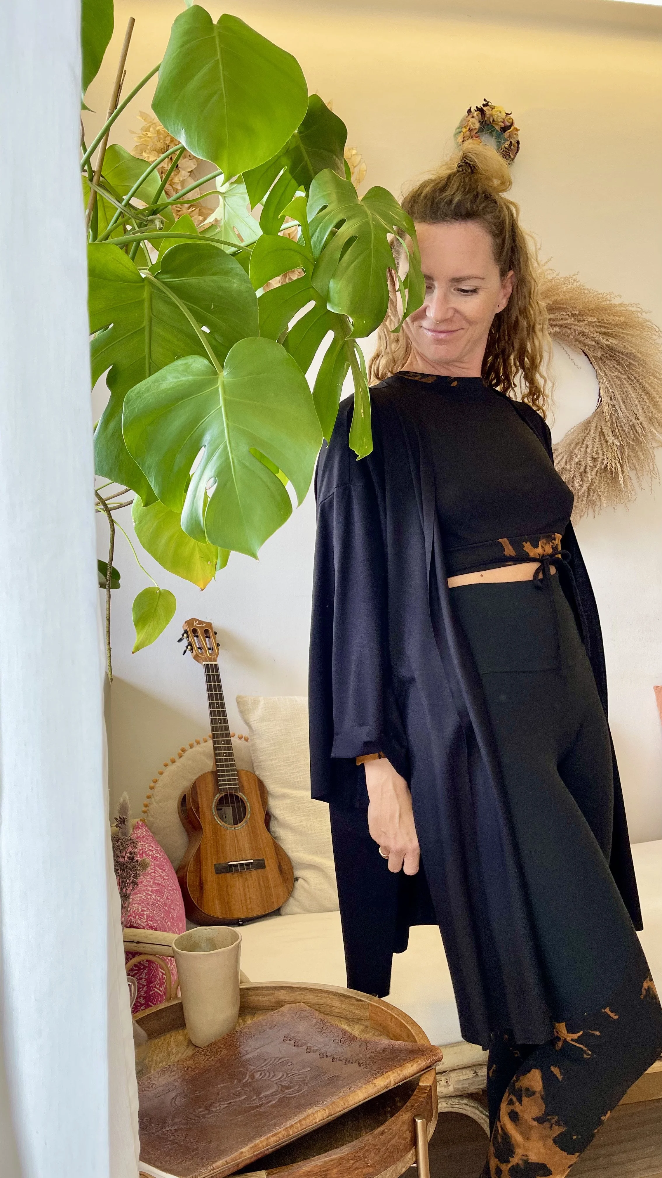 Eine Frau steht vor einer großen Monstera-Pflanze in einem Wohnzimmer, im Hintergrund eine Gitarre und Deko-Elemente.