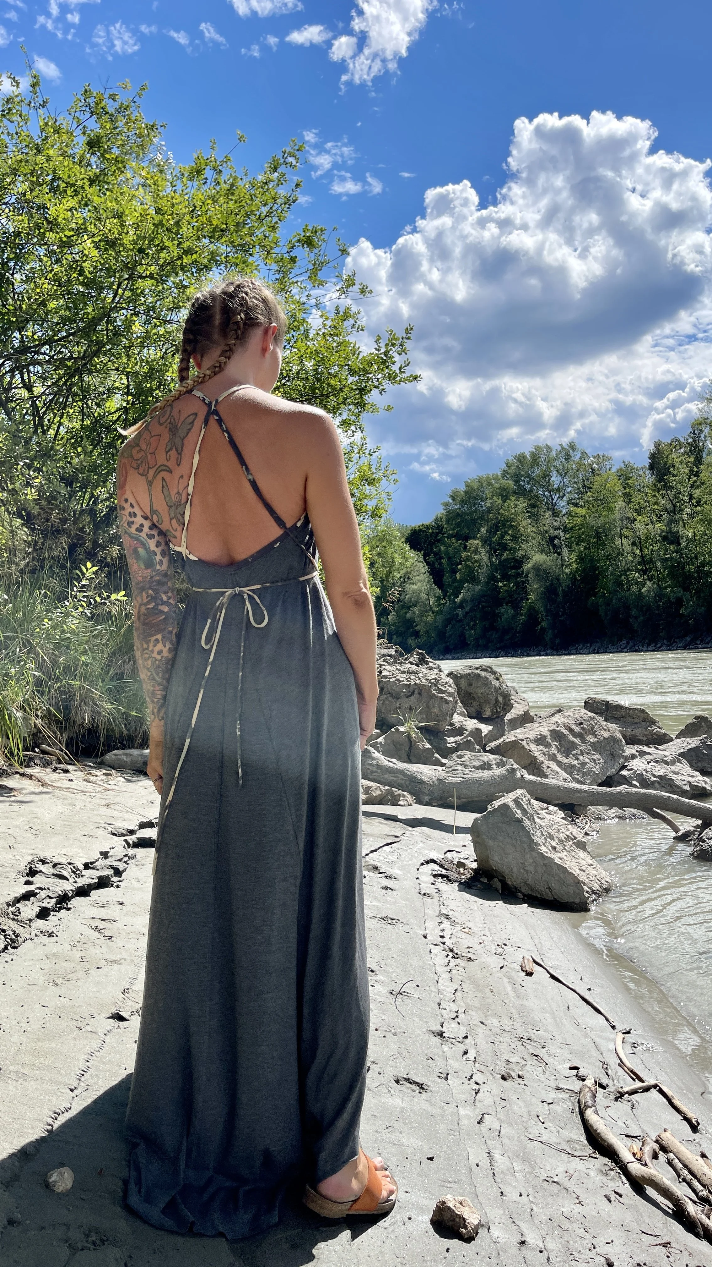 Eine Frau mit Tattoo auf dem Rücken steht am Flussufer, Blick auf die Wasser und umgeben von Bäumen, bei sonnigem Himmel mit Wolken.