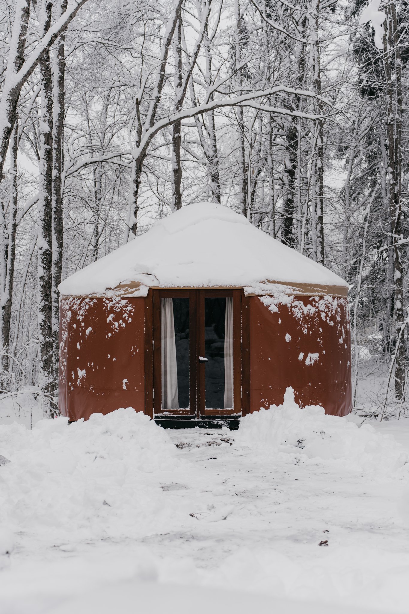 A round, brown yurt with snow on the roof, situated in a snow-covered forest with leafless trees in the background.