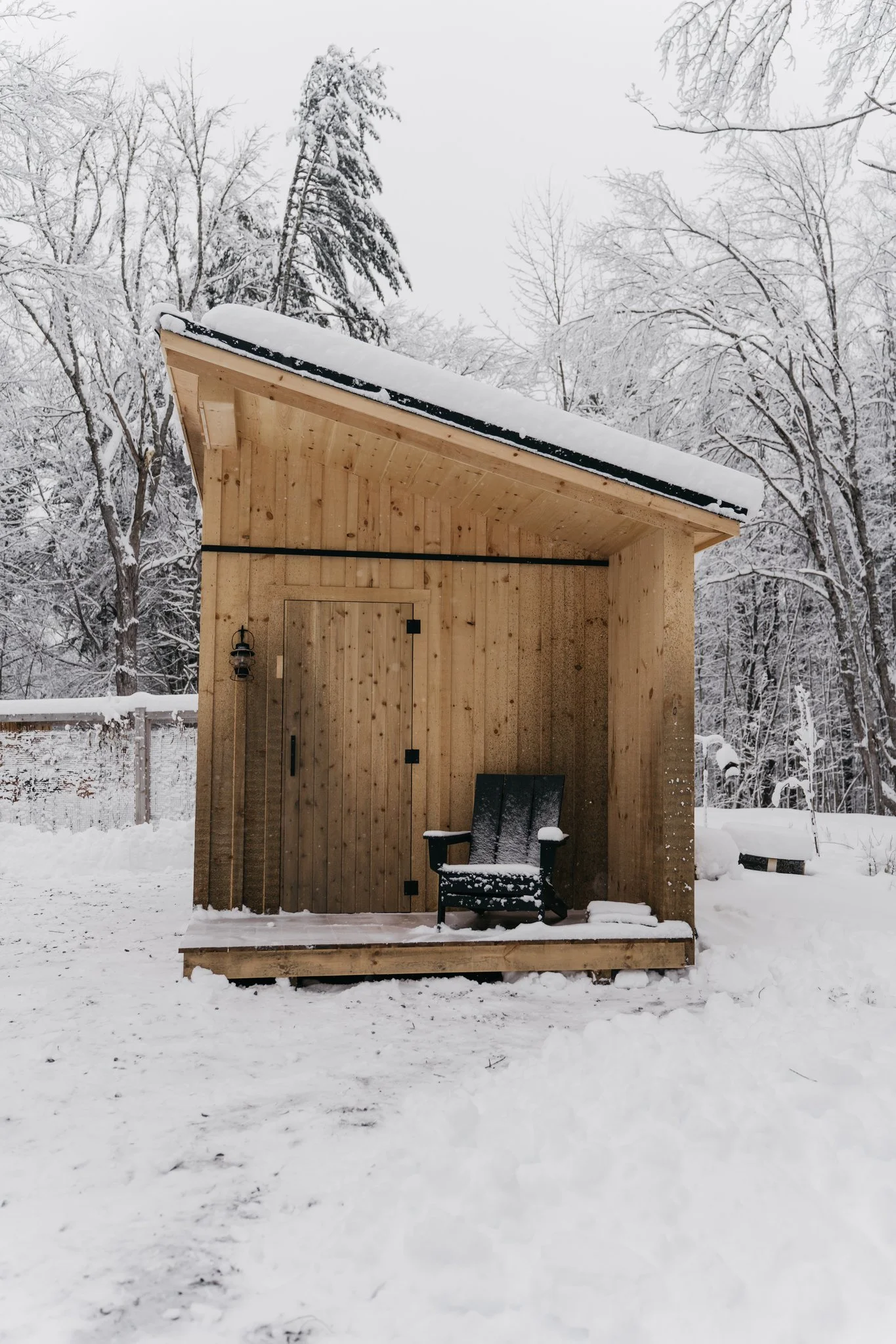 A small wooden outdoor shelter with a snow-covered roof, situated in a snowy landscape with trees in the background, and a black chair on its porch.