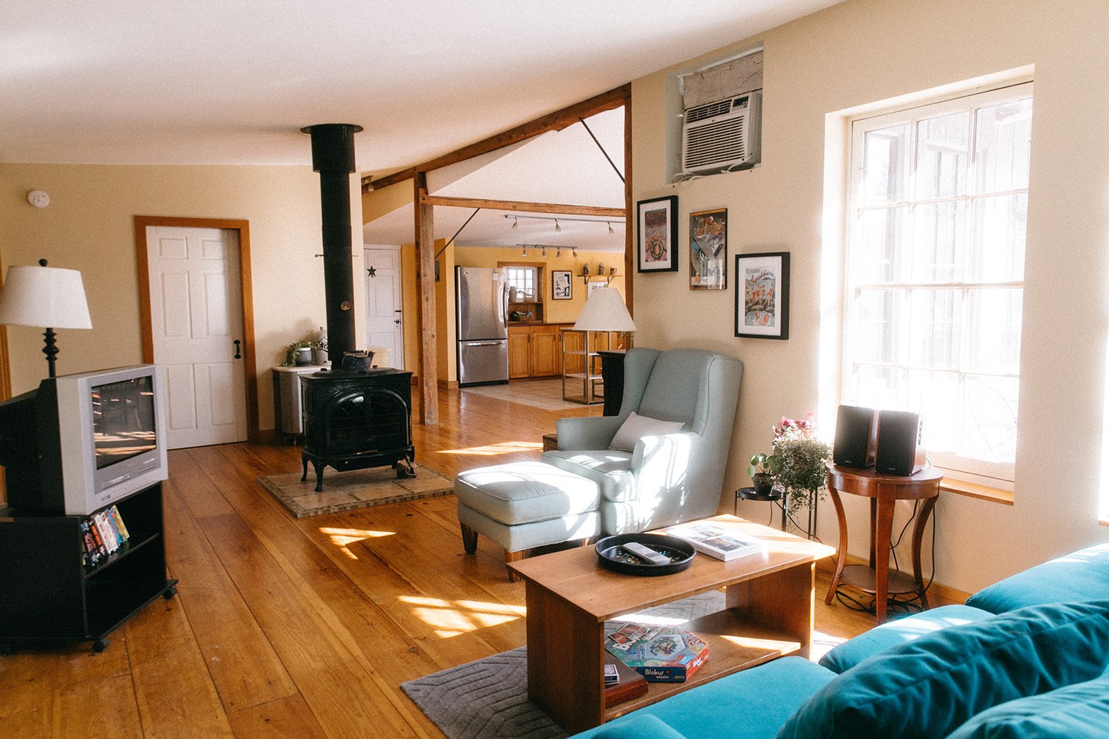 Living room with wooden flooring, light-colored walls, a TV on a small black shelf, a blue sofa, a white armchair, a wooden coffee table with magazines and a game box, a window with sunlight, and artwork on the wall. The kitchen in the background has