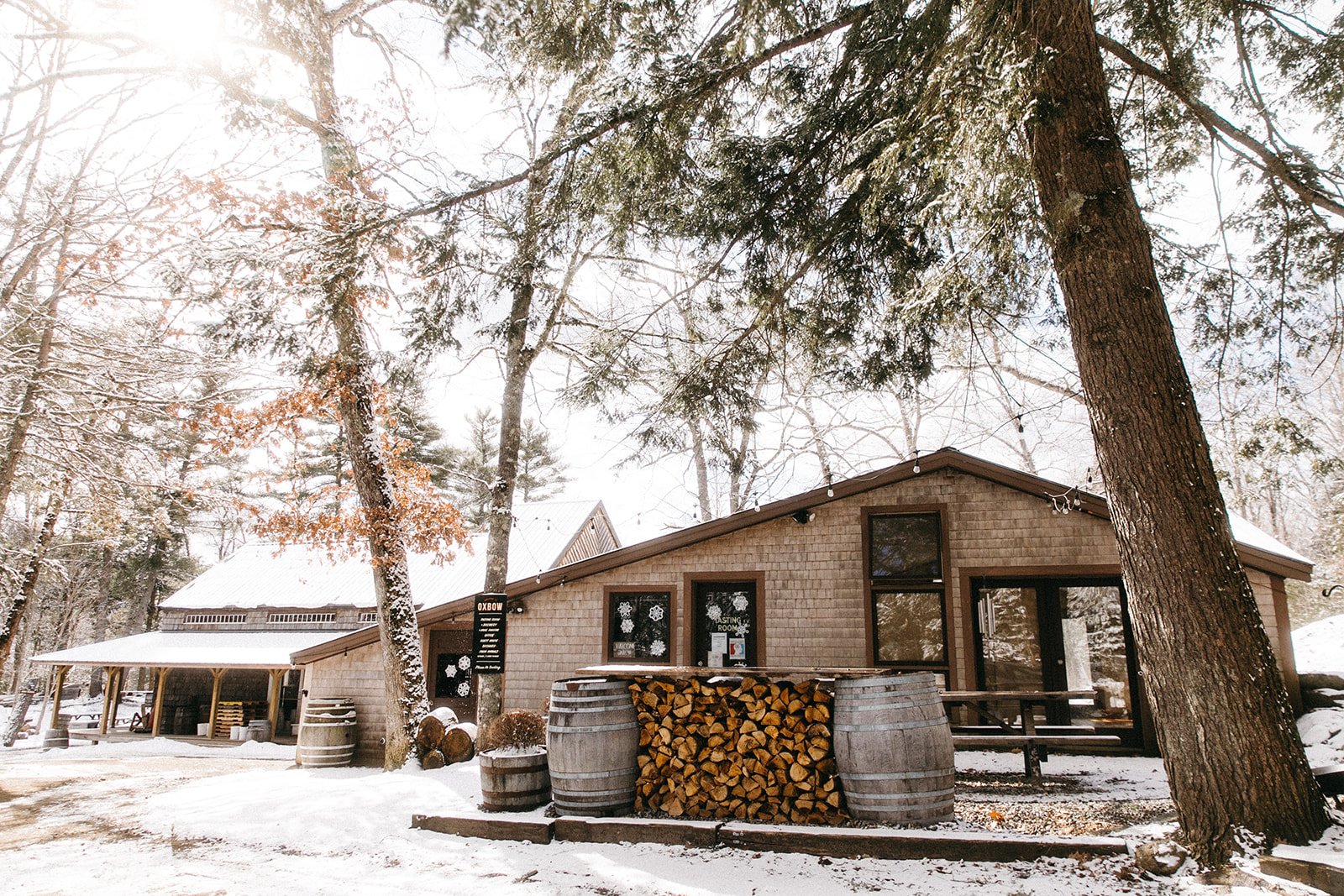 A rustic building in a snow-covered forest with stacked firewood and barrels outside, surrounded by tall trees with snow on their branches.