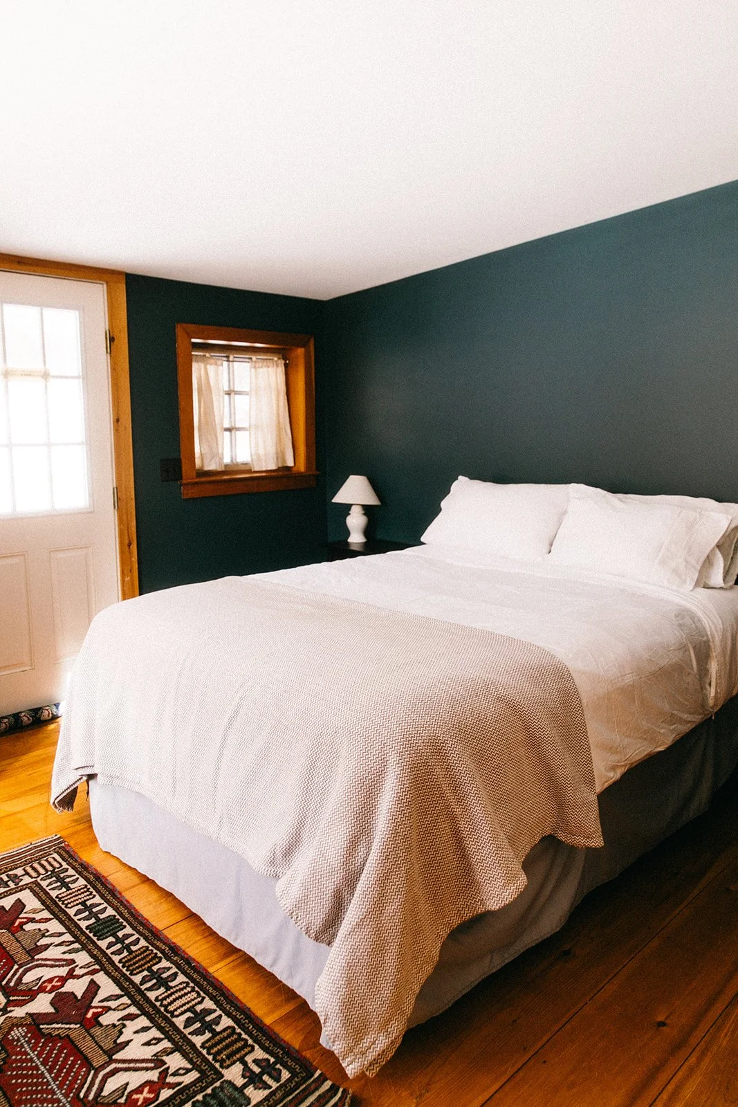 A cozy bedroom with a white bed, dark green wall, wooden accents, a small window with curtains, a door with a window, a nightstand with a white lamp, and a patterned area rug.