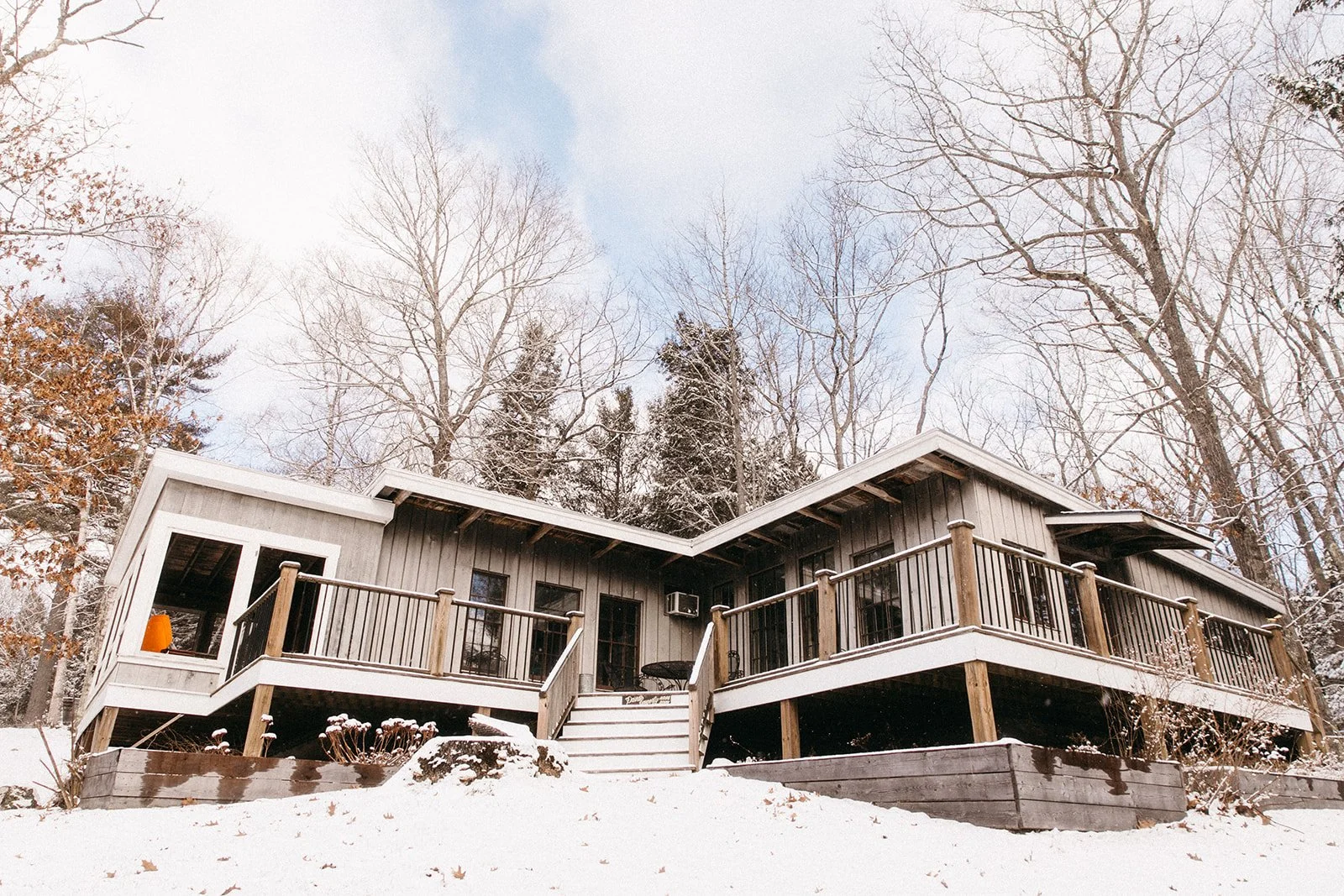 Snow-covered house with a wooden deck and railing, surrounded by leafless trees in winter.