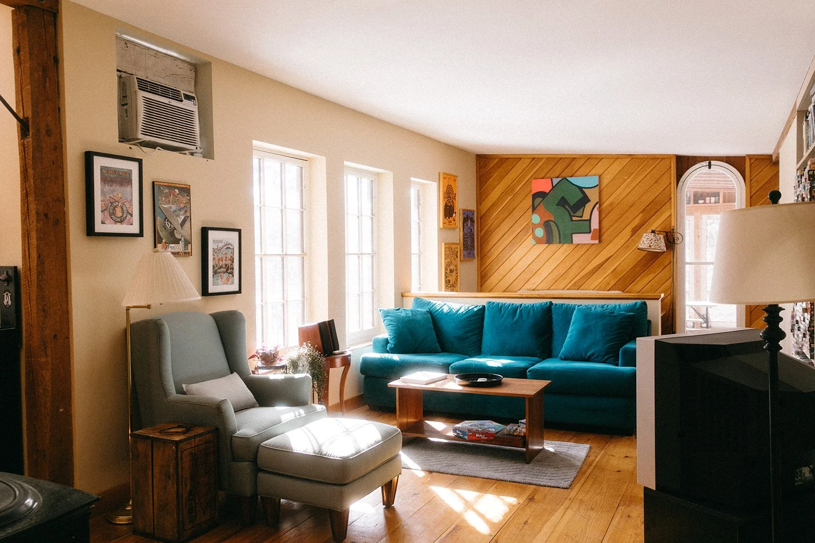 Living room with beige walls and wooden floors, featuring a light gray armchair with a white pillow, a blue couch, a wooden coffee table, various framed artwork on the walls, and large windows letting in natural light.