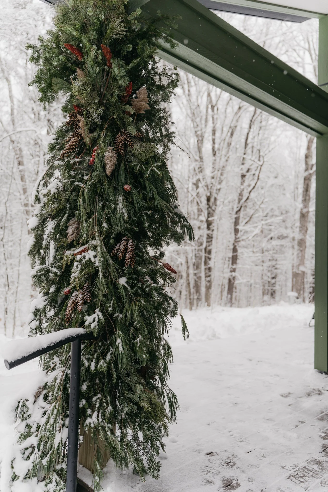 A green garland decorated with pine cones, red berries, and other pine branches, hanging outside under a green roof with snow on the ground and a snowy forest in the background.