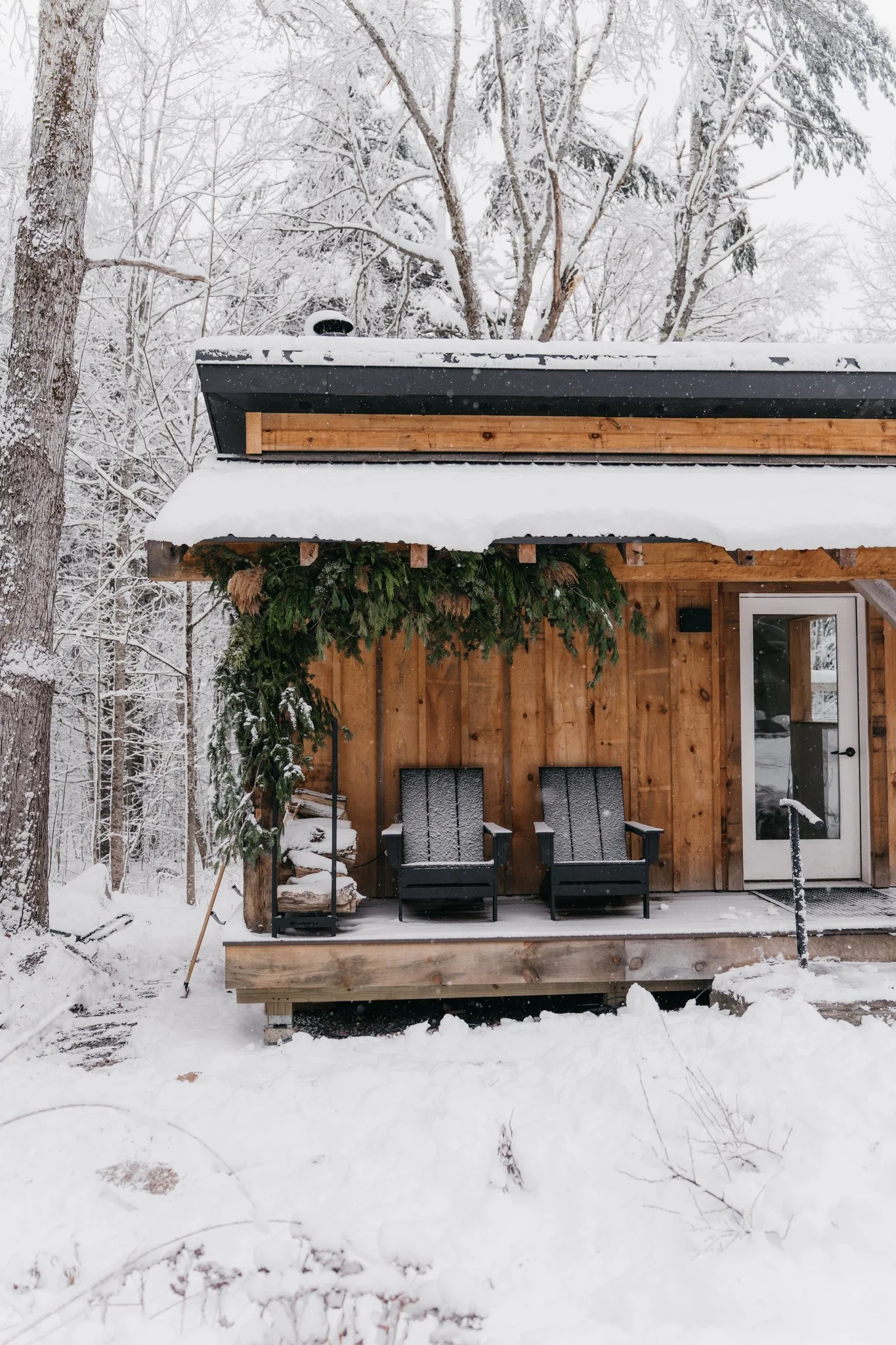 snowy wooden sauna porch with two black chairs, green holiday garland, and a glass door, surrounded by snow-covered trees.