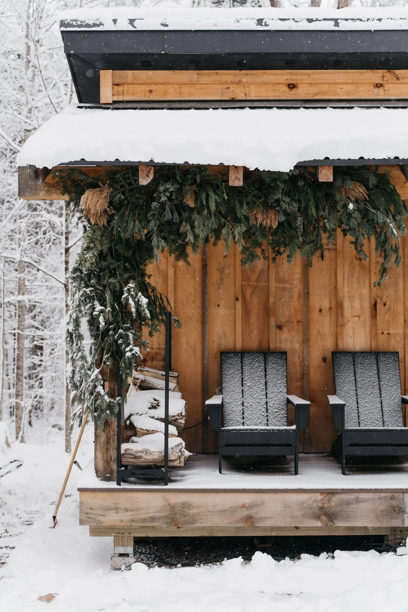 A wooden porch with two black chairs covered in snow, decorated with greenery, with a snow-covered forest in the background.