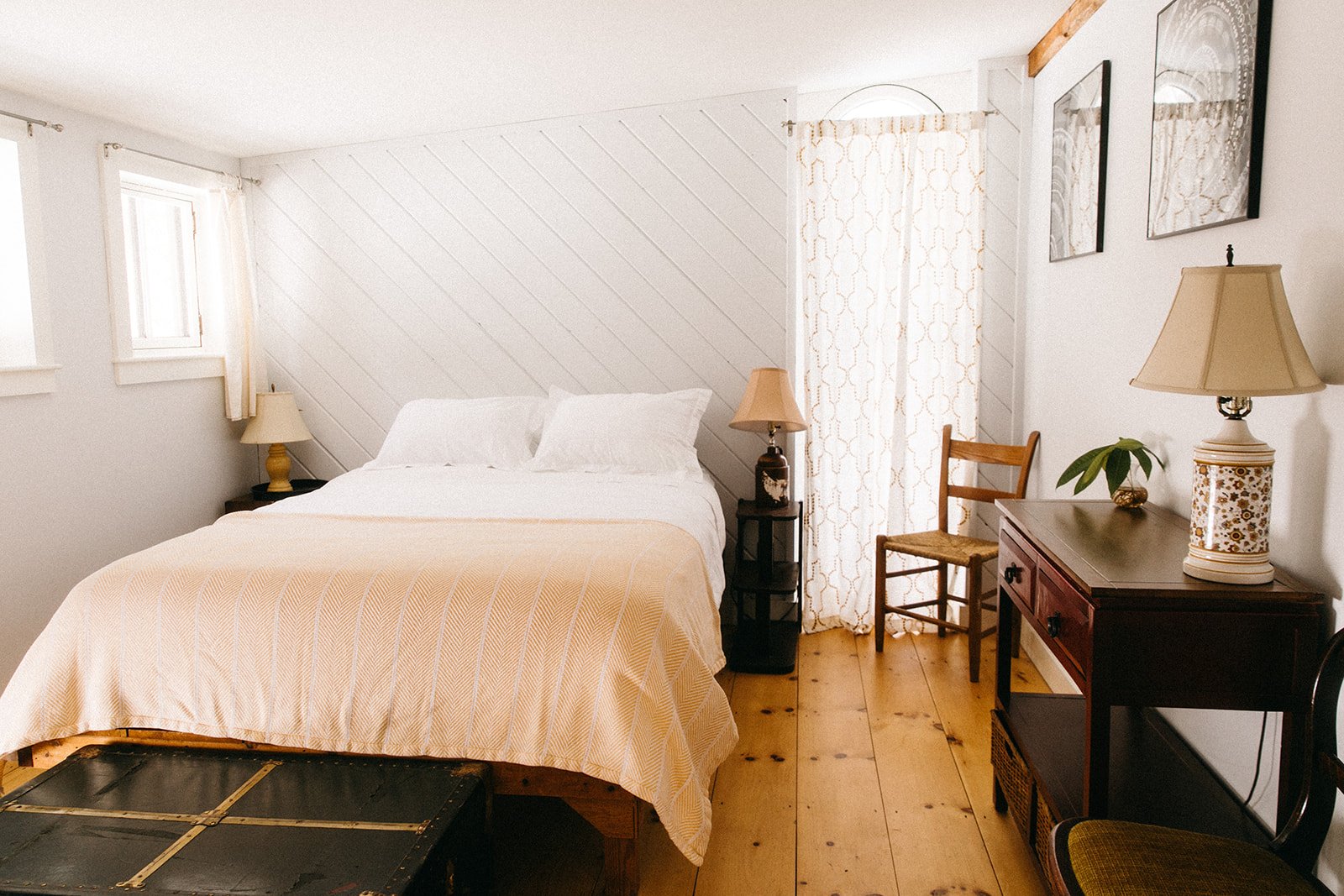 A bedroom with a neatly made bed, two bedside lamps, a wooden chair, a dark wooden dresser with a lamp and a plant, and framed artwork on white walls with wooden flooring.
