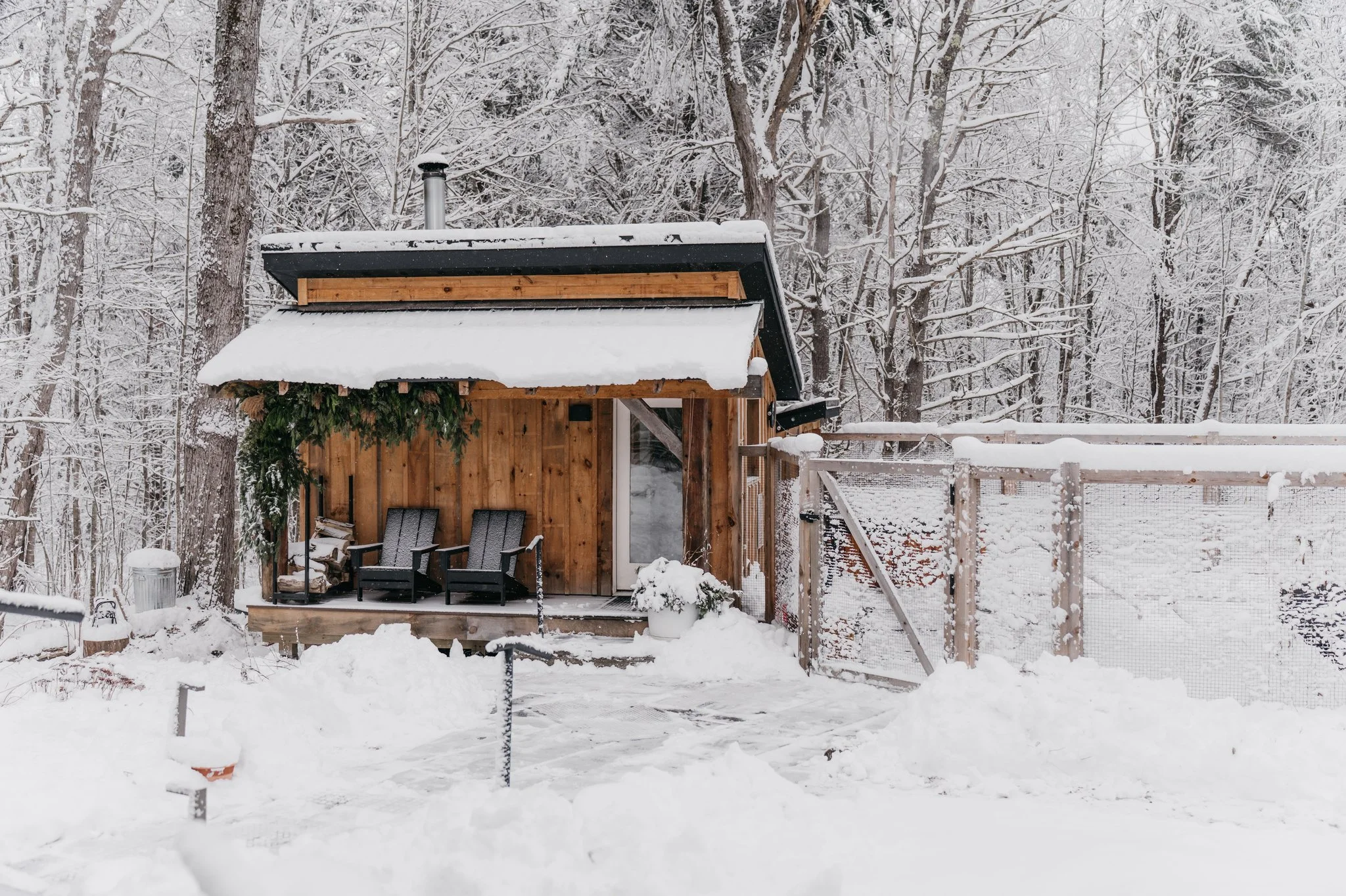 A small wooden cabin surrounded by snow, with snow-covered trees in the background. The cabin has two black chairs on a porch, a snow-covered canopy, and a potted plant with snow nearby.