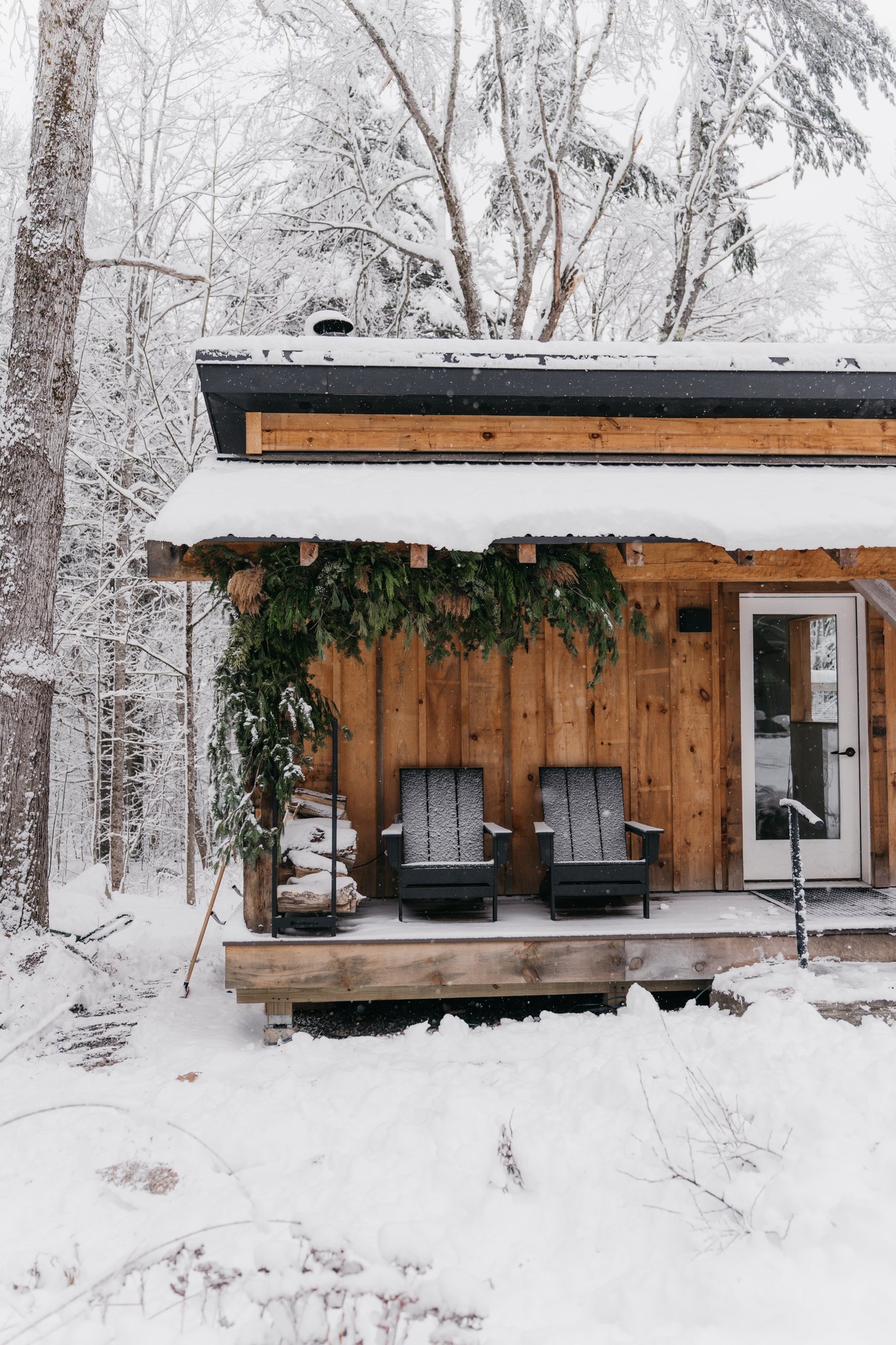 A wooden cabin or shed in a snowy landscape with two black chairs on the porch, decorated with greenery and snow.